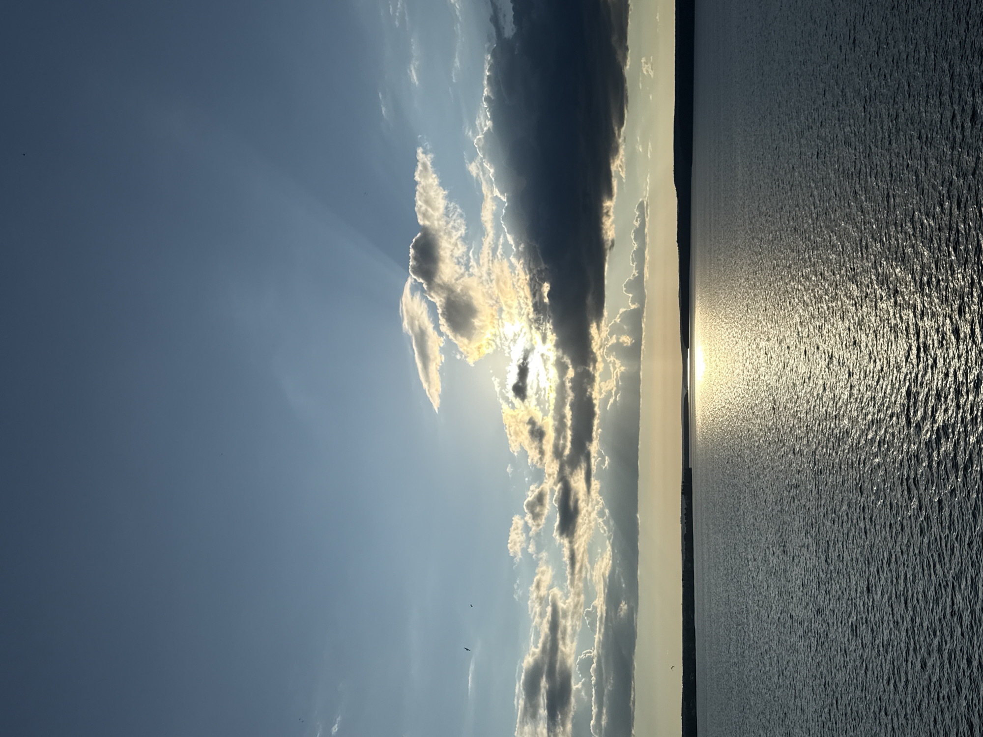 The image depicts a serene natural landscape, likely a body of water such as a lake or river, under a dynamic sky. The sun is partially obscured by clouds, casting a warm glow on the water's surface, creating a shimmering reflection. Rays of sunlight are visible piercing through the clouds, indicating it is either sunrise or sunset. The sky is a gradient of blue, with scattered clouds ranging from light and wispy to dark and ominous. A distant treeline is visible on the horizon, forming a dark silhouette against the lighter sky. The water is calm, with subtle ripples indicating gentle movement. Several small, dark specks are visible in the sky, which could be birds in flight. The scene evokes a sense of tranquility and natural beauty. The location is identified as Votkinsk, Russia, a city known for its picturesque natural surroundings.