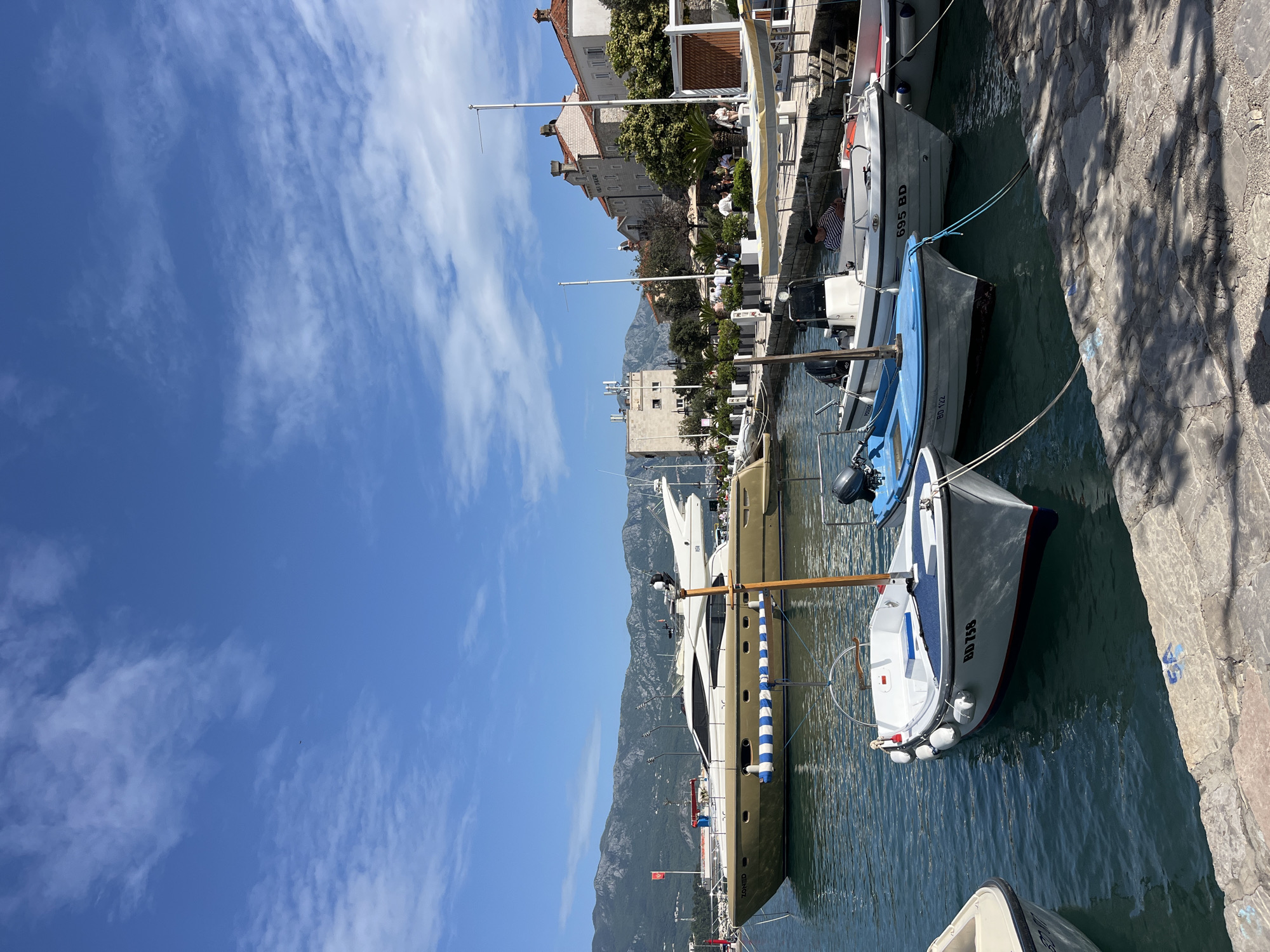The image captures a harbor scene in Budva, Montenegro. Several boats of varying sizes are docked along a concrete quay. Buildings line the harbor, with a mountainous backdrop in the distance. The sky is blue with scattered clouds. People can be seen near some of the buildings.