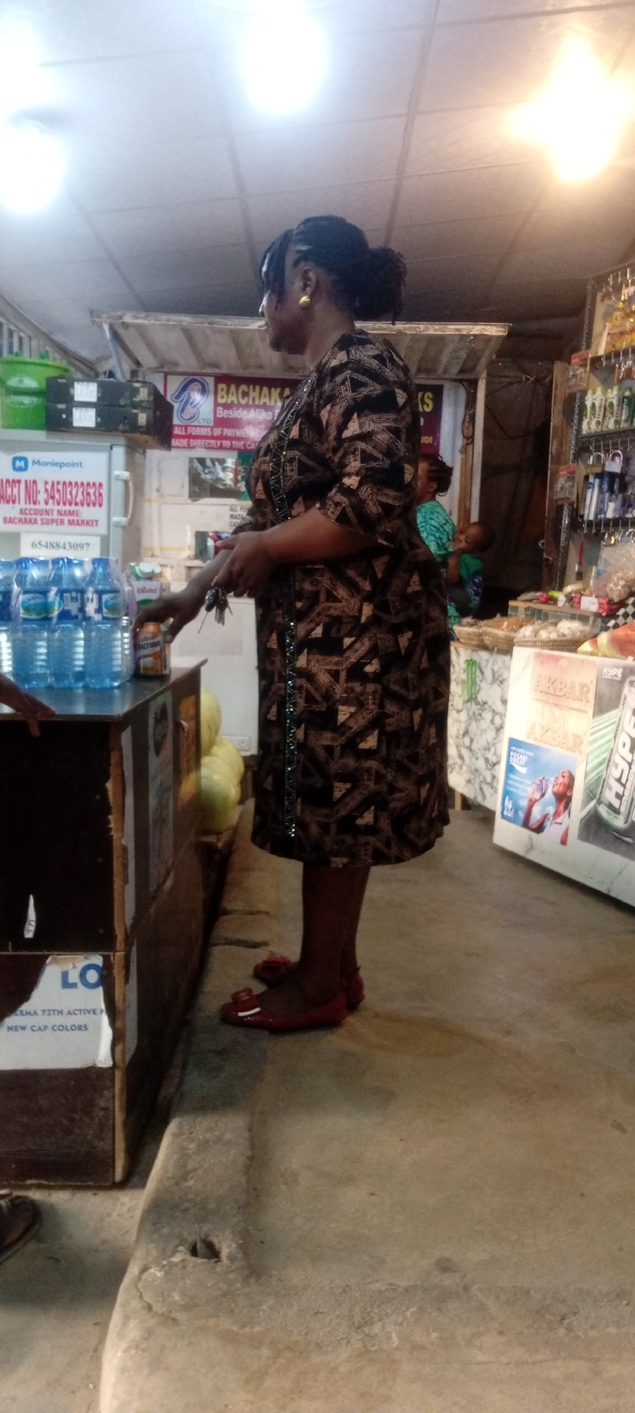 A woman with dark hair in a bun, wearing a patterned dress and red flats, stands in what appears to be a small shop in Kano, Nigeria. She is at a counter where bottled water and other items are displayed. Signage visible includes "BACHAKA", "Moniepoint," and "AKBAR" brand advertisements. The location is near Aliko. The environment is a simple commercial space with shelves stocked with various goods. Another woman and a child are visible in the background.