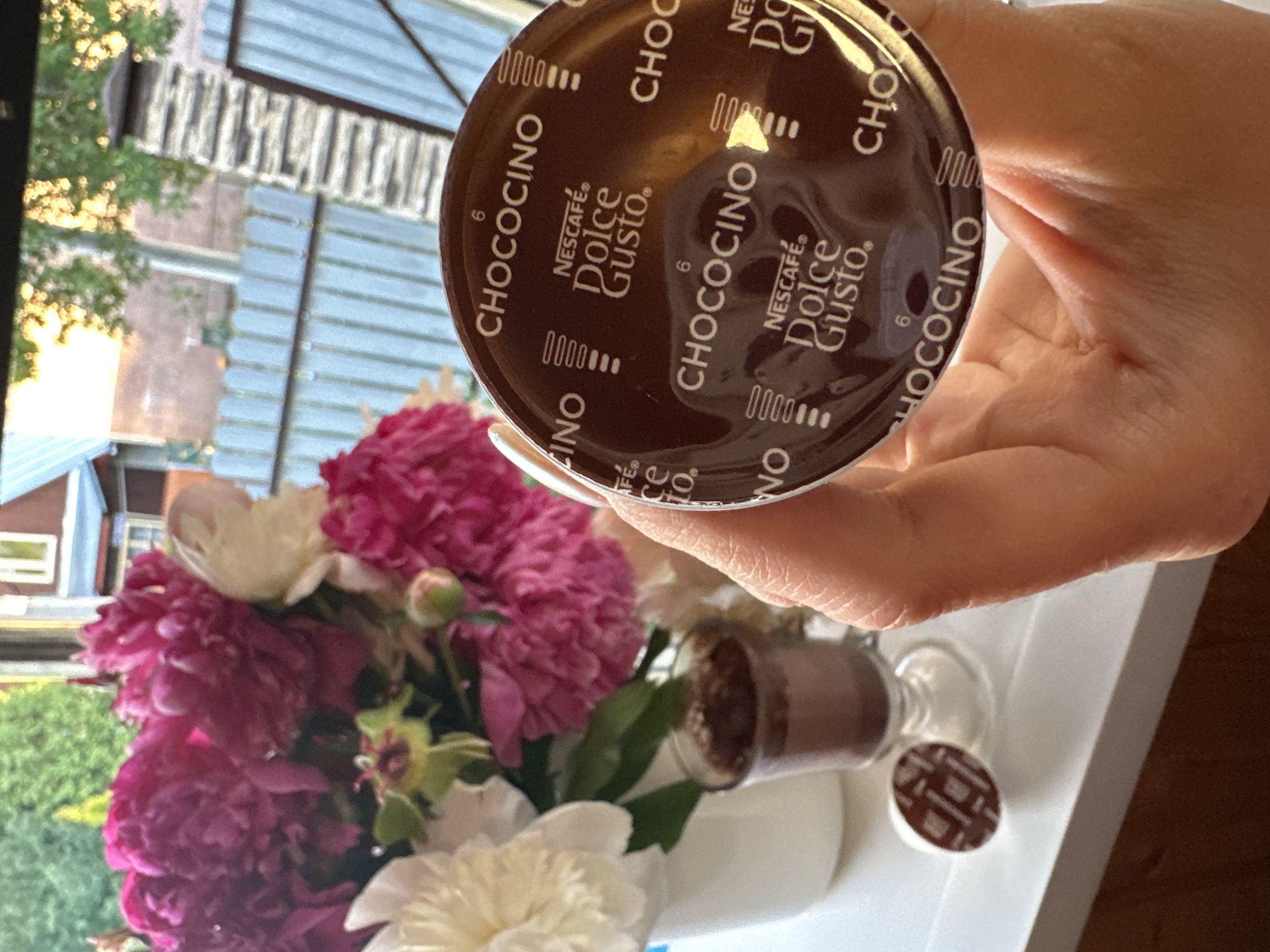 A person's hand holds a Nescafé Dolce Gusto Chococino coffee pod in front of a bouquet of pink and white peonies in a white vase. To the left of the vase is a small clear glass containing a dark brown beverage, likely hot chocolate. A single Nescafé Dolce Gusto pod, similar to the one being held, rests on the white surface in front of the glass. The background shows a blurred view of trees and a building, suggesting an outdoor or semi-outdoor setting. The presence of a garden fence and what appears to be a brick chimney suggests a residential area. This scene is consistent with a domestic setting in Nizhny Novgorod, Russia, during a time of year when peonies would be in bloom, likely late spring or early summer.