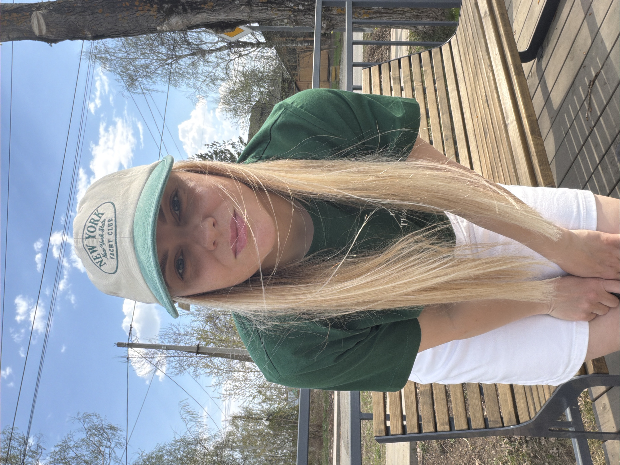 A woman with long blonde hair is seated on a wooden bench outdoors. She is wearing a green t-shirt, white shorts, and a light-colored baseball cap with the words "NEW YORK" and "YACHT CLUB" printed on it. The sky is blue with scattered white clouds, and trees are visible in the background. There are power lines overhead. The specific location within Votkinsk, Russia, is not discernible from the image.