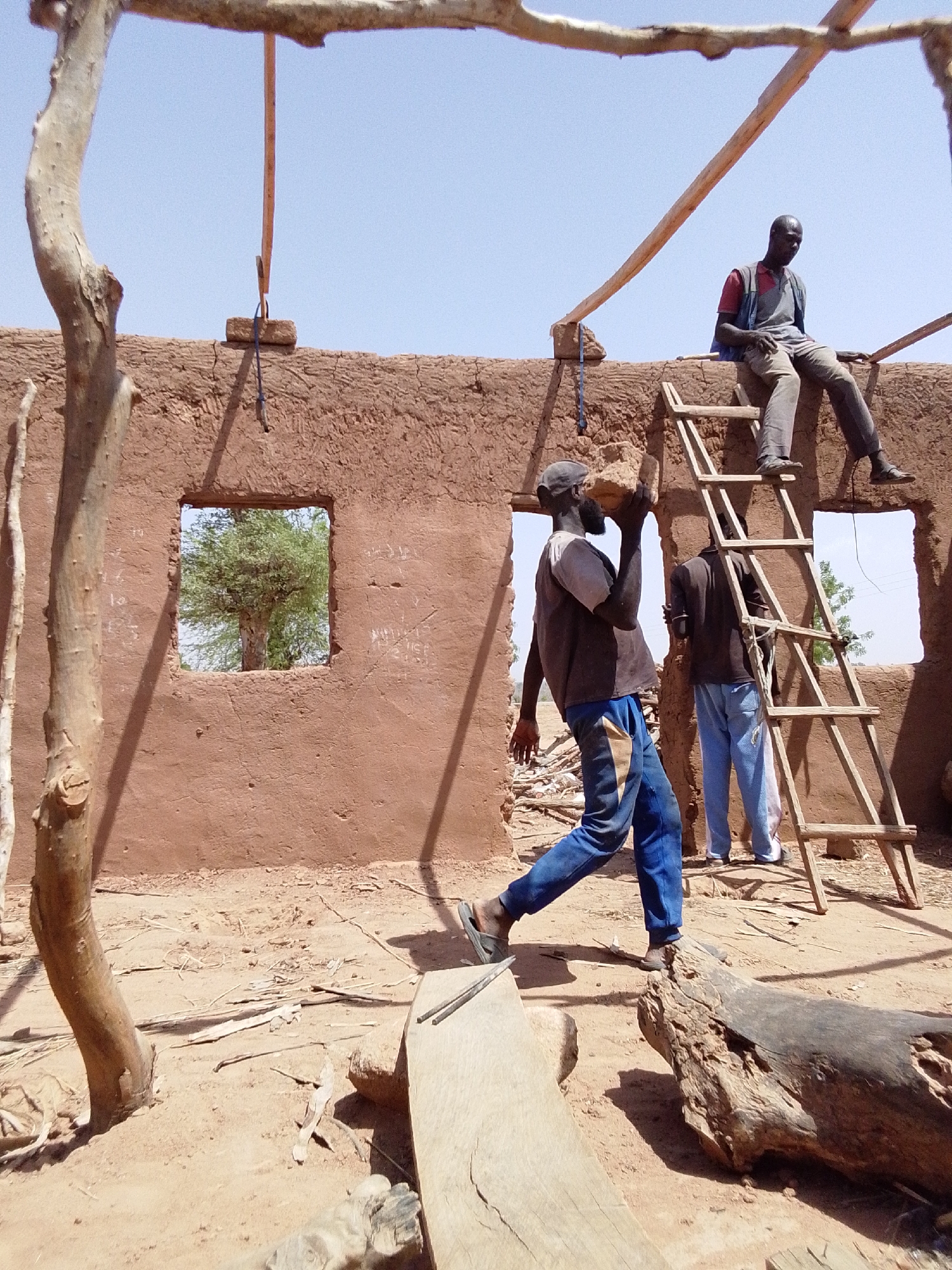 The image depicts a construction scene in Mulke Barikin Sayejo, Nigeria. A mud brick structure with window openings and wooden supports is under construction. Three men are present. One man is seated on a wooden ladder resting against the structure's wall. Another man carries a mud brick on his shoulders, while a third man stands near the ladder. The ground is dirt with scattered debris, including logs and lumber. A tree trunk and branches are visible in the foreground, framing the scene. The sky is clear and blue.