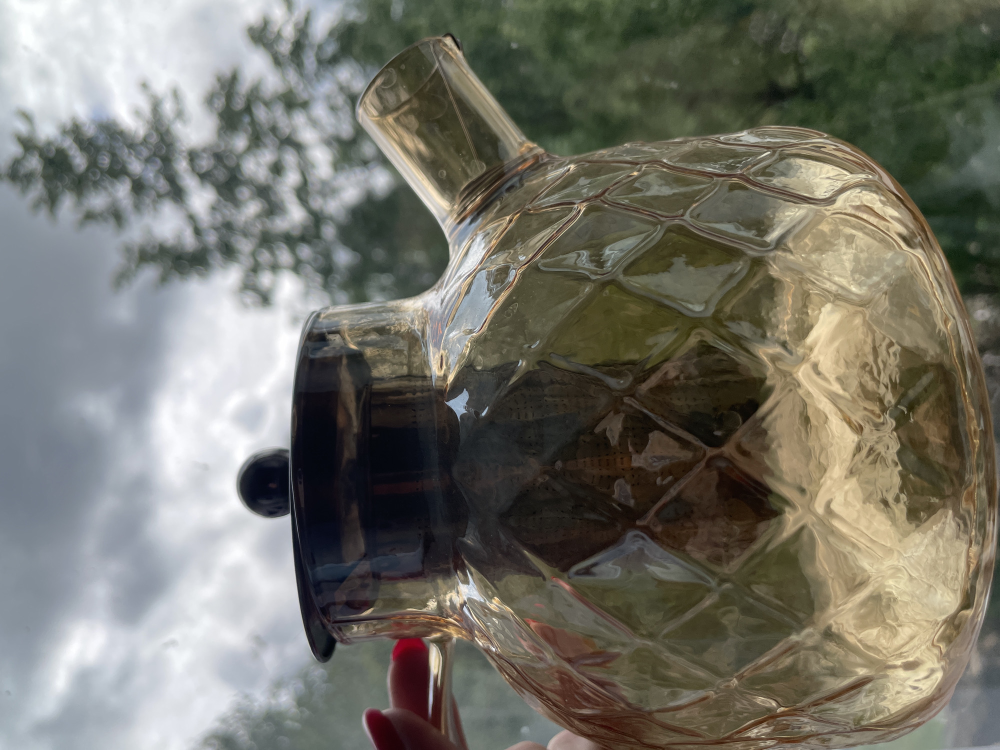 A close-up, slightly tilted, outdoor shot depicts a translucent amber glass pitcher held by a person's hand with a red fingernail. The pitcher has a unique pineapple-like texture on the body and a dark-colored lid with a spherical knob. A spout extends from the upper portion of the pitcher.
The background is blurred, showing overcast sky and green foliage, indicating a natural or park-like environment. The location is listed as Unknown, Unknown.