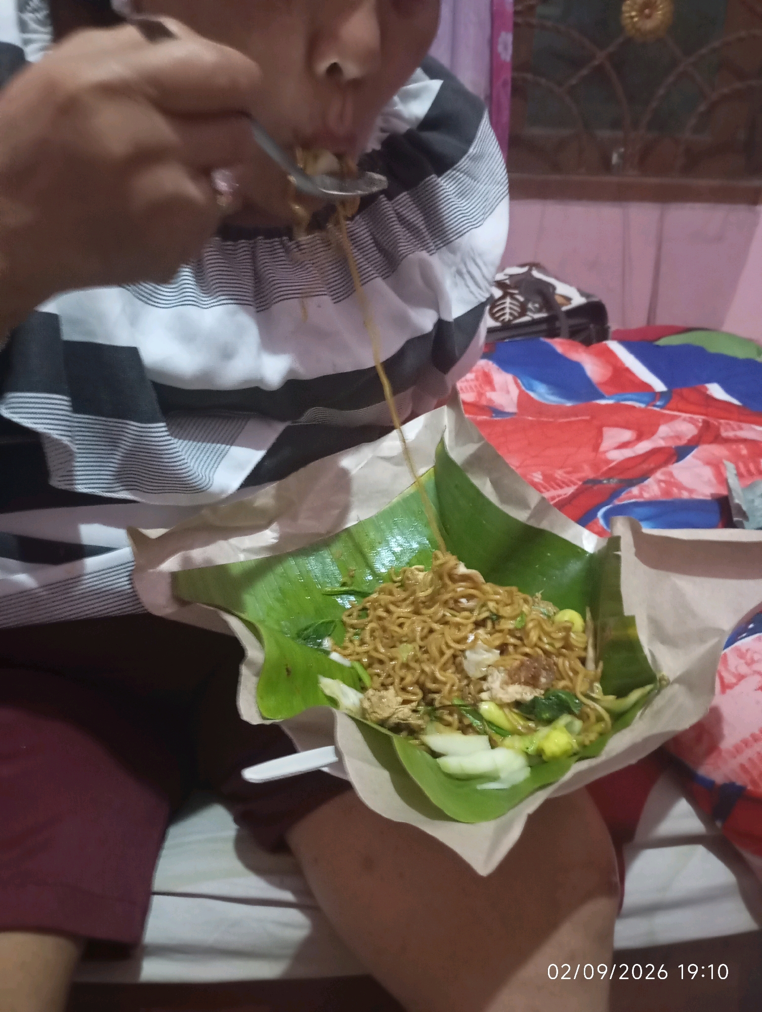 The image shows a person in Klecorejo, Indonesia, eating a meal of noodles served on a banana leaf. The person is wearing a striped shirt and maroon pants. The food is presented in a disposable container lined with brown paper and a banana leaf. The noodles appear to be stir-fried with vegetables and some protein. The person is using a spoon to eat the noodles. The background shows a bed with a colorful, patterned comforter and a glimpse of a window with a decorative grate. The timestamp at the bottom right of the image indicates the date and time as 02/09/2026 19:10.