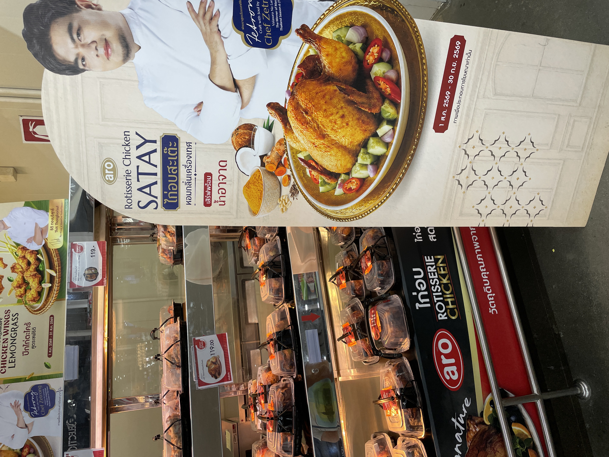 The image shows a display of rotisserie chicken in a store, likely a supermarket or deli, in Pattaya City, Thailand. A large promotional sign features a chef, Chef Jetrong, next to a whole rotisserie chicken presented on a golden platter with sliced cucumber, red onion, and chili. The sign also highlights "Rotisserie Chicken SATAY" and mentions ingredients like lemongrass and turmeric, suggesting a Thai flavor profile. Below the main sign, a red banner displays dates in Thai numerals, likely indicating a promotional period. In the background, refrigerated display cases are filled with various packaged food items, including what appear to be chicken products. The overall environment suggests a commercial setting focused on food sales.