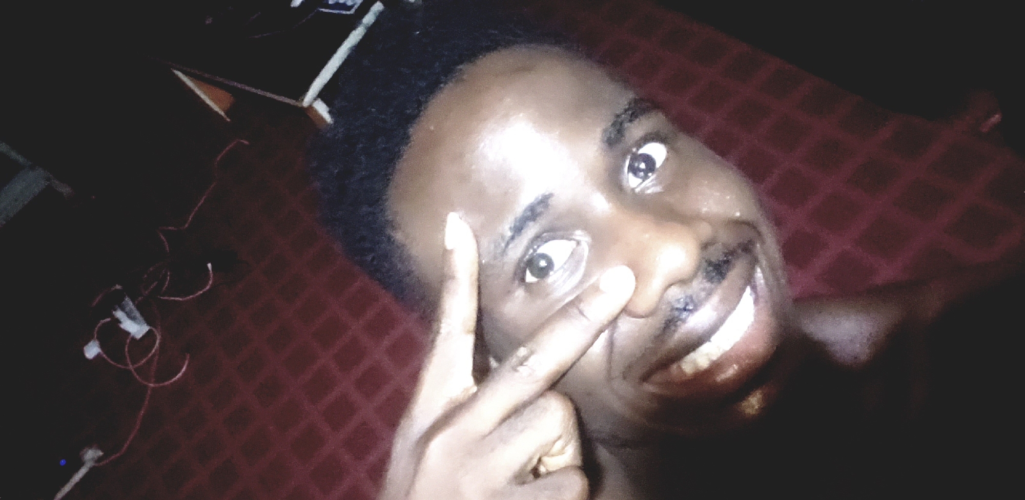 An African man is taking a selfie. The man's left index finger is pointed towards his left eye, and he is smiling. The background appears to be a patterned, reddish surface, possibly a bedspread. In the top left corner, there's a glimpse of what looks like electronic equipment with wires. It is hard to tell where in Narayi, Nigeria this image was taken due to the background information.