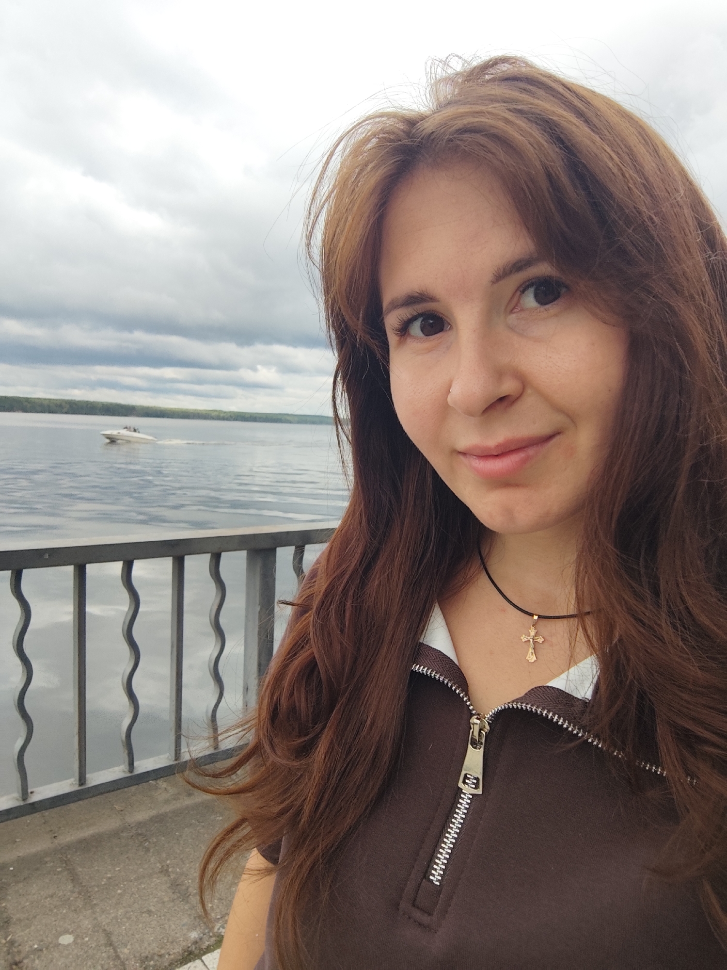 A woman with long, auburn hair takes a selfie outdoors. She is wearing a brown zippered sweatshirt with a white collar and a gold cross necklace. In the background, there is a body of water, a grey metal railing, and a cloudy sky. A white boat is visible on the water in the distance. The location is likely the embankment along the Kama River in Chaikovsky, Russia, as indicated by the presence of the river and the general landscape.