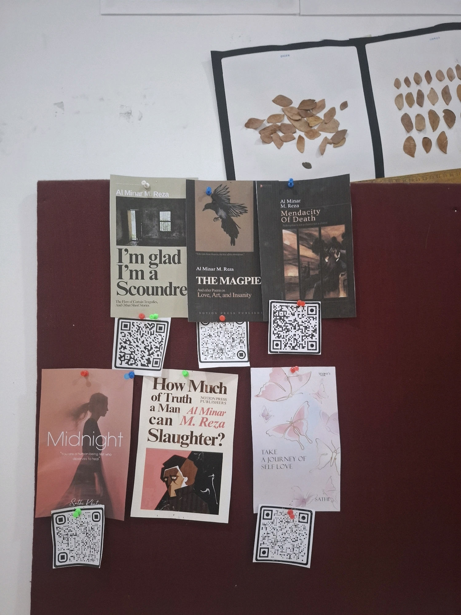 The image depicts a bulletin board with several books and printed materials pinned to a dark red surface. The top portion of the board has two white sheets of paper with dried leaves arranged on them. Below these, four book covers are pinned: "I'm glad I'm a Scoundre" by Al Minar M. Reza, "The Magpie" and other poems by Al Minar M. Reza, "Mendacity of Death" by Al Minar M. Reza, and "Midnight" by Sathi Khatri. Additionally, a flyer titled "How Much of Truth can a Man Slaughter?" by Al Minar M. Reza is pinned to the board. A smaller card with butterflies and the text "TAKE A JOURNEY OF SELF LOVE" is also visible. Several QR codes are attached to the board, presumably linked to the books or authors. The background appears to be a white wall. The scene is indoors, with artificial lighting. There are no people visible in the image. The overall context suggests a display of literary works. The image does not provide any specific indicators of the city of Barasat, India, or any other geographical location.