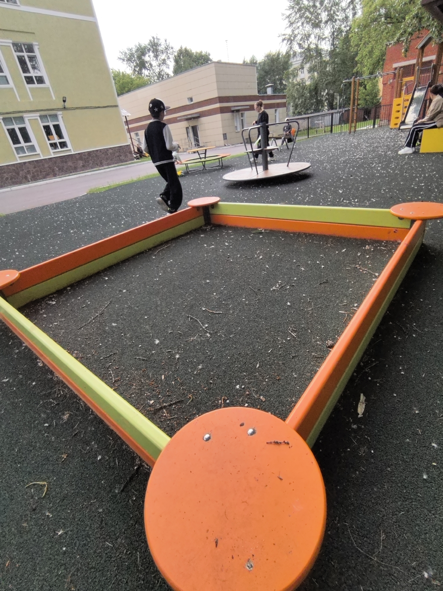 This daytime image depicts a children's playground featuring various equipment and individuals under an overcast sky. The foreground is prominently occupied by a low, triangular border structure, crafted from alternating bright orange and lime green beams, with circular orange platforms at each corner. The entire playground area is covered with dark grey rubberized safety flooring, visibly speckled with small, light-colored debris, possibly seeds or pollen.
Several people are present in the scene. To the left, a young person, wearing a black and white jacket, black pants, and a black baseball cap adorned with white stars, is captured walking carefully along the top edge of the orange and green border, facing away from the viewer. In the mid-ground, another individual in a black long-sleeved shirt and glasses is standing near a circular merry-go-round, which has a brown wooden top and a black base with handrails. Further to the right, a third person, dressed in a light brown top and dark pants, is seated on a piece of yellow playground equipment, which appears to be part of a slide or a specialized seat, situated next to a red brick building.
The background features several buildings: a light yellow multi-story building with numerous white-framed windows on the far left, a light brown building with distinctive horizontal dark brown stripes in the center, and a red brick building on the right. Green trees with dense foliage are interspersed around the playground and behind the structures. The lighting is soft and diffused, indicating an overcast day. No visible text is present on any of the playground equipment, buildings, or other elements within the frame.