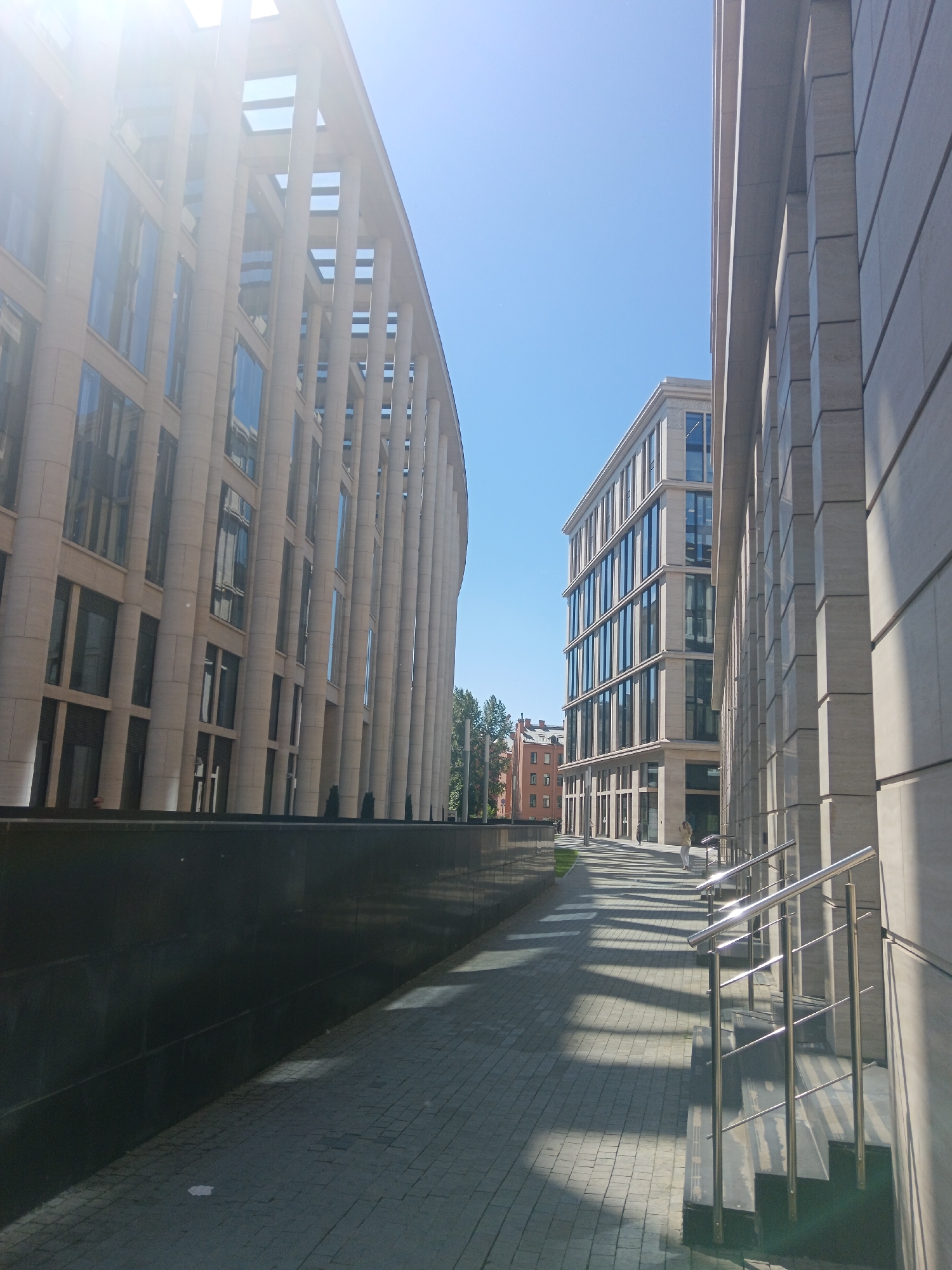 This is a sunny, eye-level, wide shot of modern architecture in an urban environment. The scene is dominated by two large buildings. The building on the left features a curved facade with many tall, slender columns and large glass windows, creating a sense of grandeur. The building on the right is a rectangular structure with a similar facade of stone and glass. Between the two buildings, a paved walkway with a patterned brick surface leads towards the buildings. There are staircases with modern stainless steel railings leading up to an entrance on the right. In the background, more buildings and some trees are visible. The sky is a clear, bright blue, and strong sunlight casts long shadows across the walkway, indicating it's likely midday. There are a few individuals walking in the distance, one on the walkway and another near the entrance of the building on the right. The overall impression is one of clean, contemporary design in a well-maintained urban setting.