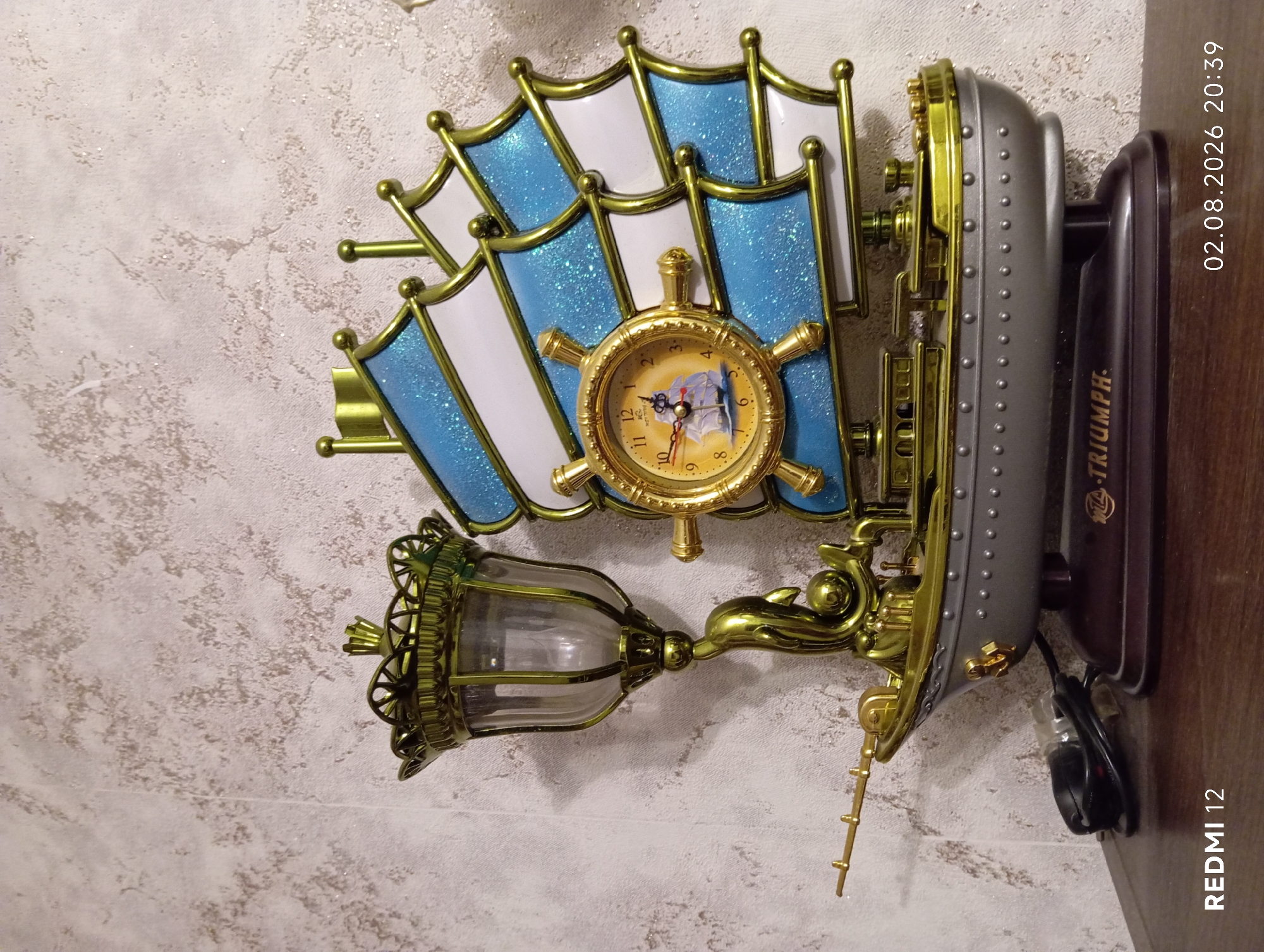 The image depicts a decorative item shaped like a ship, likely a clock or a music box, on a wooden surface against a textured wallpaper. The ship is predominantly gold and silver, with blue and white sails. It features a steering wheel with a clock face, a lamp with a glass shade supported by a golden dolphin, and a fishing rod extending from the bow. The ship has the inscription "TRIUMPH" on its base. The timestamp "02.08.2026 20:39" and "REDMI 12" are overlaid on the image, indicating the capture details. There are no people or animals present in the image. The environment appears to be indoors, possibly a home setting. The location in the city of Oryol, Russia is not visually discernible from the image itself; this information would need to be provided externally.