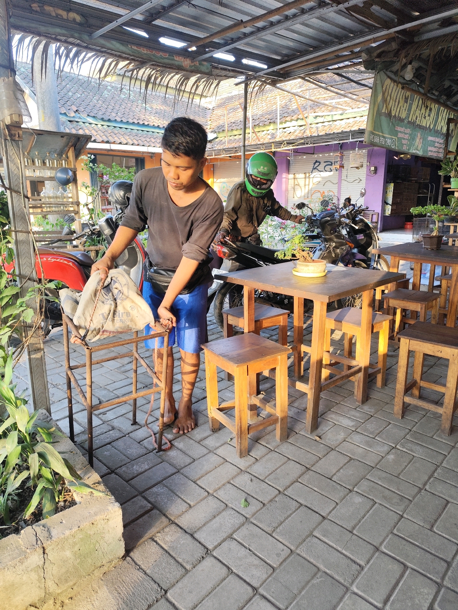 In Bambang Lipuro, Indonesia, two individuals are present under a covered outdoor seating area. The person in the foreground, a male with short dark hair, wears a black long-sleeved shirt and blue shorts. He is standing and holding a beige cloth bag with rope handles over a rustic metal stand. A black fanny pack is worn around his waist. To his left, a red motorcycle is parked. In the background, a second person, wearing a green helmet and dark clothing, is seated on a dark motorcycle, which is positioned in front of a building with purple walls and an awning. The ground is paved with rectangular gray bricks. Wooden tables and stools are arranged in the seating area. Green plants are visible in pots and along the edge of the seating area. The scene appears to be an outdoor cafe or warung.