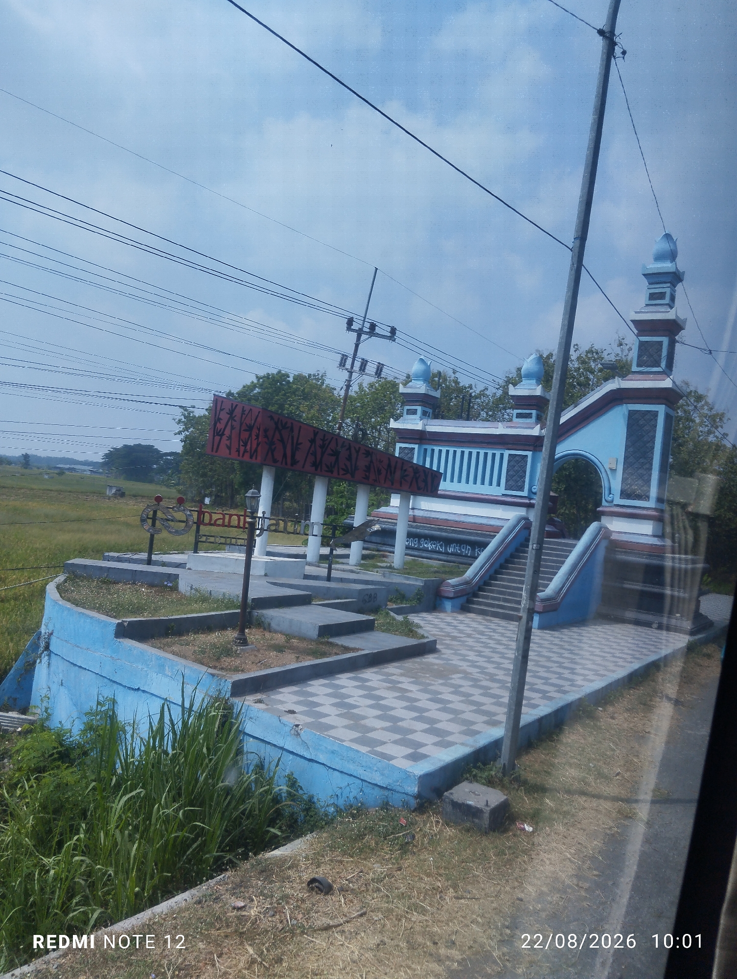 The image captures an outdoor scene in Baluk, Indonesia, featuring a blue and white archway structure adorned with decorative pillars and a patterned roof. A red sign with a bamboo motif is prominently displayed on a white pillar. In the foreground, a checkered tiled pathway leads up to the archway, with blue-painted walls and steps on either side. The area is surrounded by dry grass and some greenery. In the background, a field stretches out under a cloudy sky. Power lines crisscross the sky above the scene. The lower left corner of the image shows "REDMI NOTE 12" and the lower right corner displays "22/08/2026 10:01", indicating the device and timestamp of the photo.