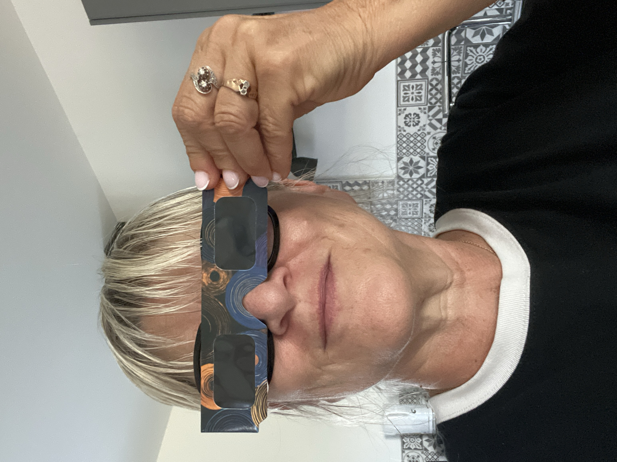 A woman with blonde hair and fair skin is holding a pair of eclipse glasses to her face. She is wearing a black t-shirt with a white collar. Her right hand is visible, adorned with two rings, and her fingernails are painted pink. The background appears to be an indoor setting with patterned tiles on the wall. The location is identified as Chambœuf, France.