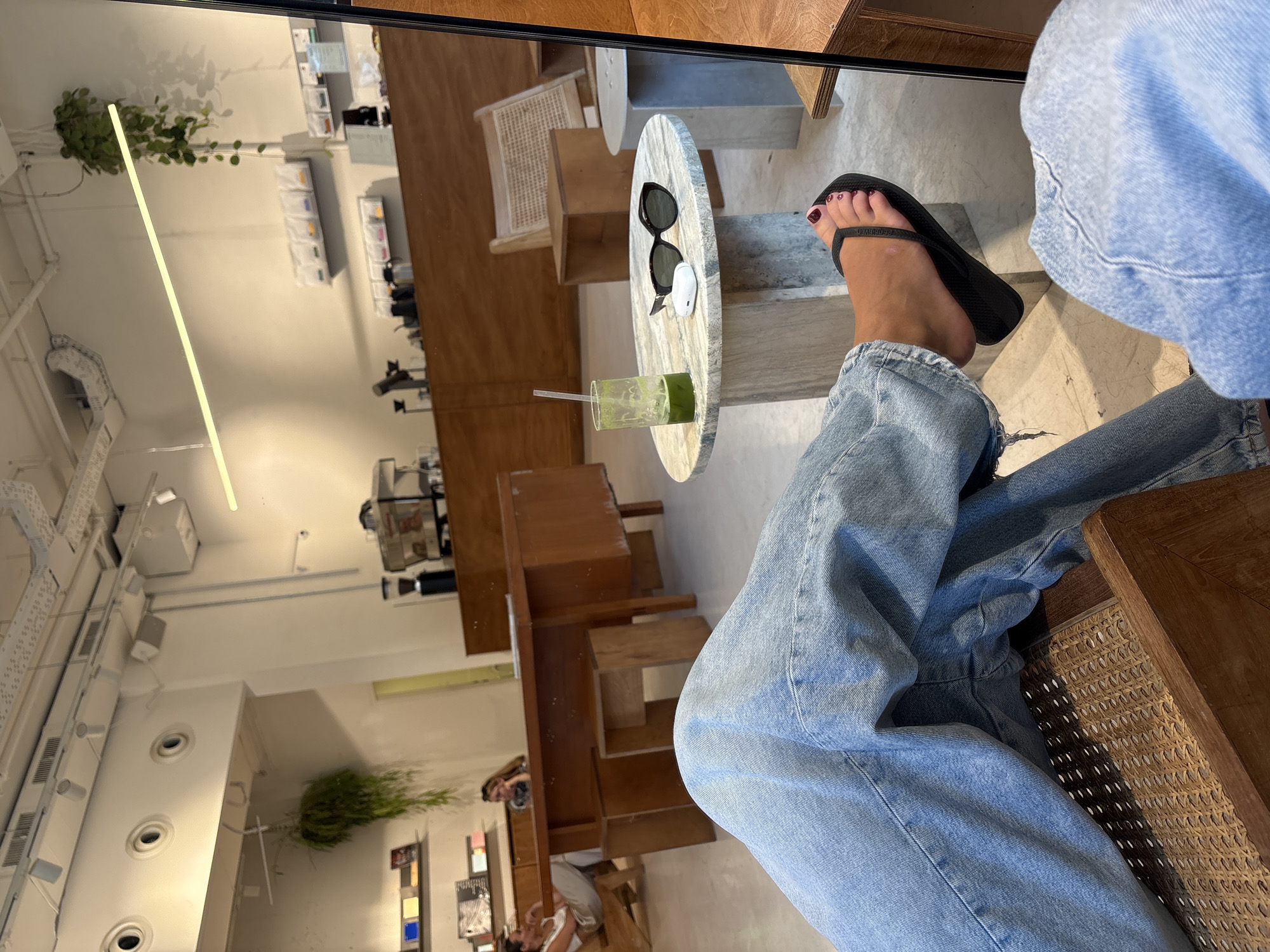 The image depicts the interior of a bright, modern cafe in Tel Aviv, Israel. The perspective is from a seated person's viewpoint, focusing on their lower legs clad in light-wash denim jeans and black flip-flops. In the foreground, a wicker chair is visible. To the right, a marble-topped circular table holds a glass of green liquid with a straw, a pair of black sunglasses, and white wireless earbuds. In the mid-ground, a long wooden counter with coffee machines is present, and further back, two women are seated at a wooden table, one appearing to be in conversation. Shelves displaying books and small items line the wall behind them. The cafe's ceiling features exposed conduits and track lighting, with a hanging plant adding a touch of greenery. The overall ambiance is relaxed and casual, suggesting a typical cafe scene in the city.