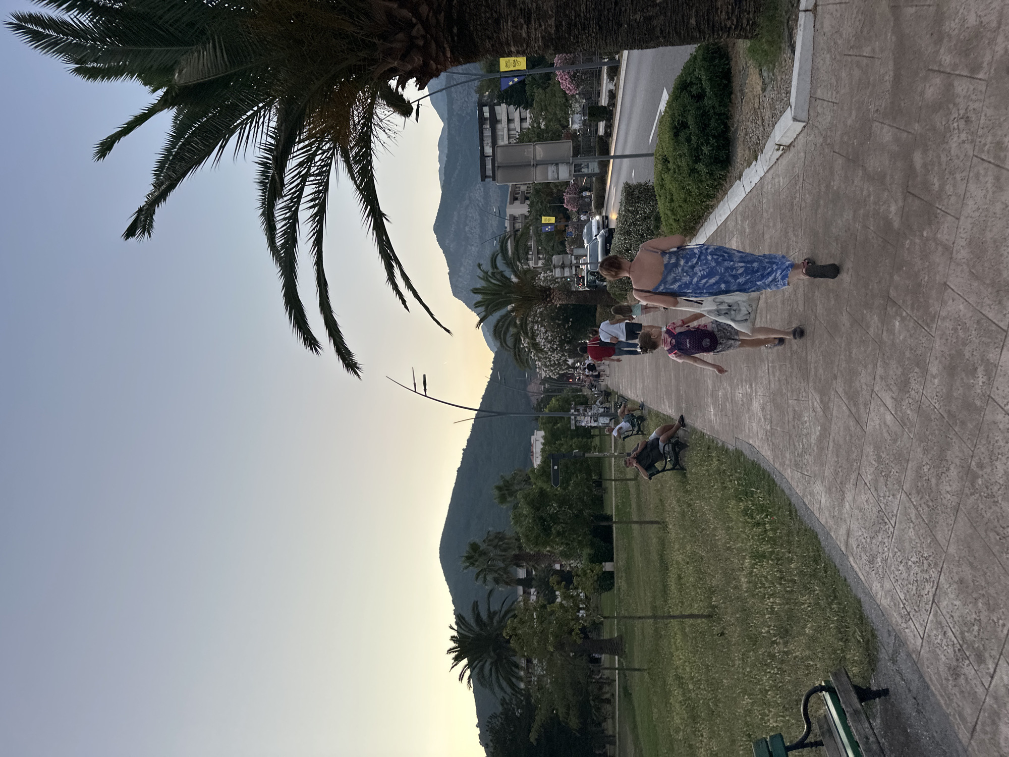 In Budva, Montenegro, a woman in a blue floral dress and a child with a backpack walk along a paved walkway. They are facing away from the camera, moving towards a road with passing cars. On the left side of the walkway is a grassy area with benches, some occupied by people sitting. Palm trees and other greenery are visible throughout the scene, with a large, tree-covered mountain in the background. The sky is a pale gradient of blue and yellow, suggesting it is either dawn or dusk.