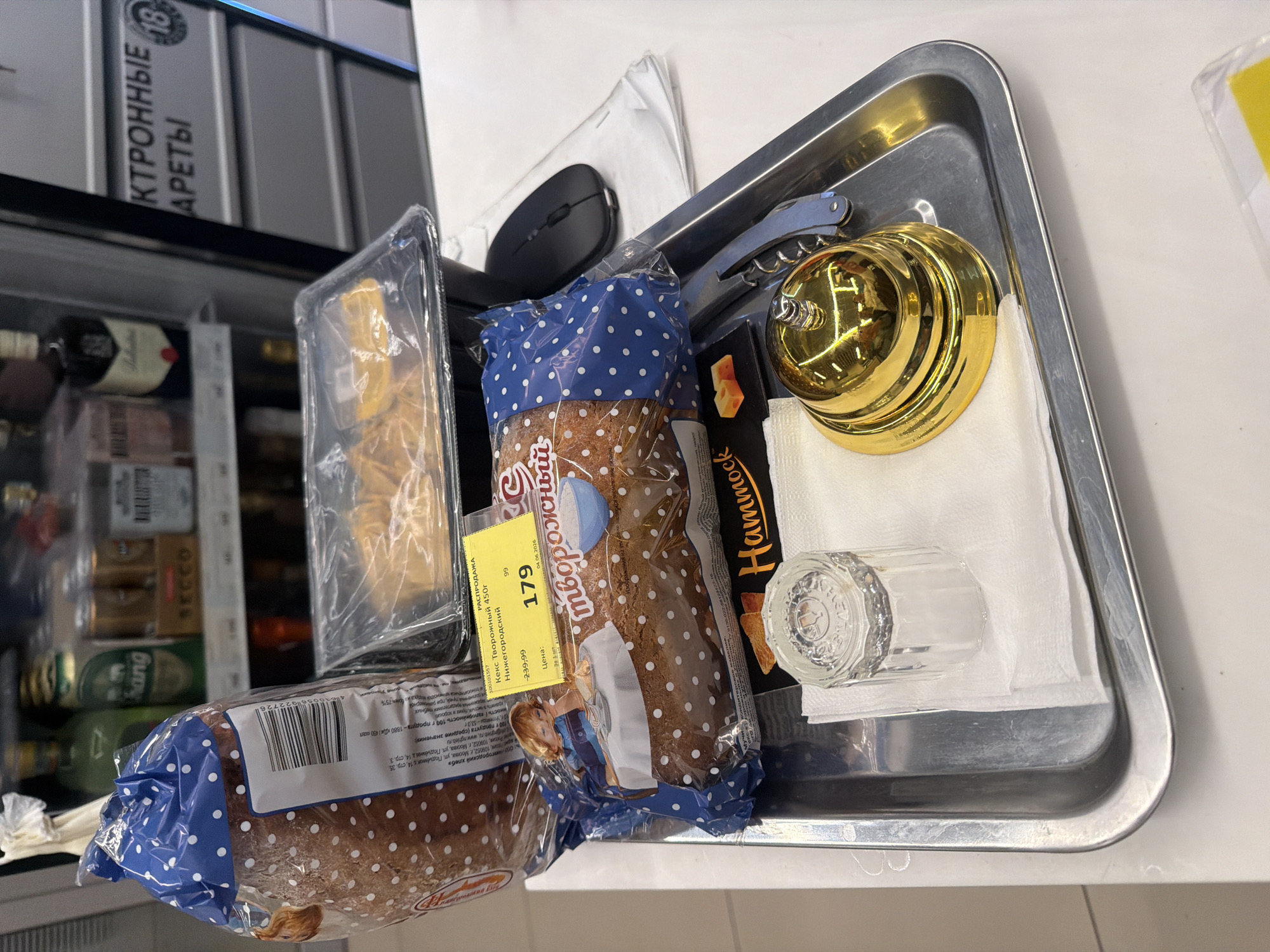 The image captures an indoor scene, likely in a hotel room or a small shop area, featuring a metal tray alongside a refrigerated display case. No people are visible.
On the shiny metal tray, several items are neatly arranged: a black computer mouse, a silver corkscrew/bottle opener, a golden call bell, a folded white napkin, and a clear, ribbed glass. Beside the glass, a dark package, possibly a snack, is visible with the word "Hammock" on it.
Adjacent to the tray are various packaged goods. Two packages of "Кекс Творожный 450г" (Curd Cake 450g), from "Нижегородский" (Nizhny Novgorod), are prominently displayed. One cake package has a yellow price tag indicating a "РАСПРОДАЖА" (SALE) price of 179 (down from 239.99), with an expiration date of 04.06.2026. The packaging features a blue polka-dot design and a cartoon image of a girl. A barcode (4600694022726) and nutritional information (1880 kJ / 450 kcal per 100g, 53g carbohydrates) are visible on the back of one package. Next to these, a clear plastic container holds what appears to be pastries or other baked goods.
In the background, a refrigerated display case is partially visible, containing various beverages and other items. Bottles of wine (one with a "Ballantine's" tag) and cans of beer, including "Chang" brand, are discernible. Above the fridge, a sign clearly states "ЭЛЕКТРОННЫЕ СИГАРЕТЫ" (Electronic Cigarettes) with an "18" age restriction, indicating the sale of such products. The visible address on the cake package mentions Moscow, Russia.