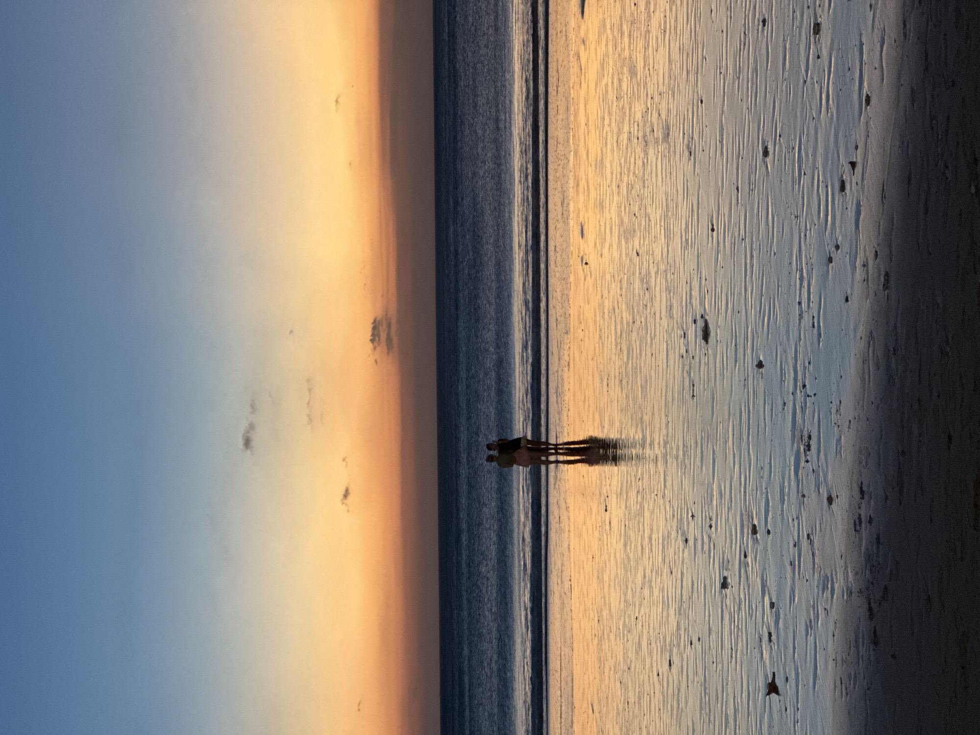 The media file captures a serene beach landscape during sunset or sunrise in Kuta Selatan, Indonesia, though the image itself is presented in a vertical orientation.
The sky, occupying the upper portion of the scene, displays a dramatic gradient of colors, transitioning from a clear, soft blue on the left to a vibrant, warm orange and yellow towards the right, indicative of the golden hour. A few small, dark, wispy clouds are scattered within the warmer hues of the sky.
A horizontal band of dark blue ocean water stretches across the mid-ground, separating the sky from the expansive wet sand of the beach in the foreground. The water appears calm, with a subtle line marking the gentle interface where waves meet the shore.
Two human figures, appearing as silhouettes, stand together at the water's edge, slightly to the right of the center. They face towards the horizon, seemingly observing the vista. Their elongated, distinct shadows stretch across the reflective, wet sand towards the viewer, emphasizing the low angle of the sun. The sand itself exhibits intricate ripple patterns and textures, reflecting the warm sky colors, with scattered small, dark specks.
The scene evokes a profound sense of peace, beauty, and contemplation, characteristic of a tranquil tropical beach at dusk or dawn. There is no visible text in the image.