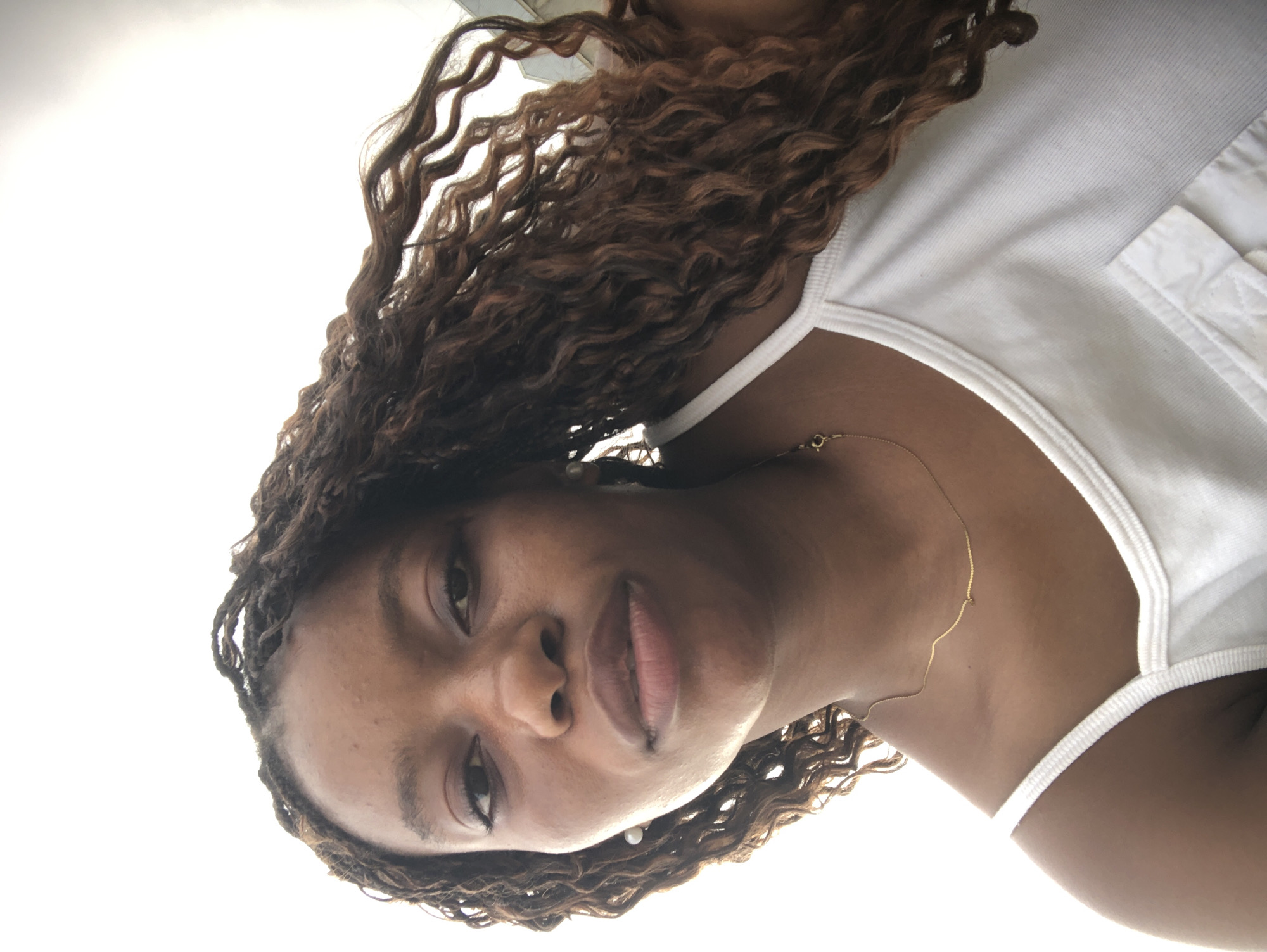 A medium-shot, downward-angled photograph depicts a woman with dark skin, brown eyes, and dark brown, wavy hair. She is wearing a white tank top and a thin gold necklace. The background is a bright, indistinct white. There is no discernible information in the image to place the scene in Kashere, Nigeria, or to infer specific actions or interactions.