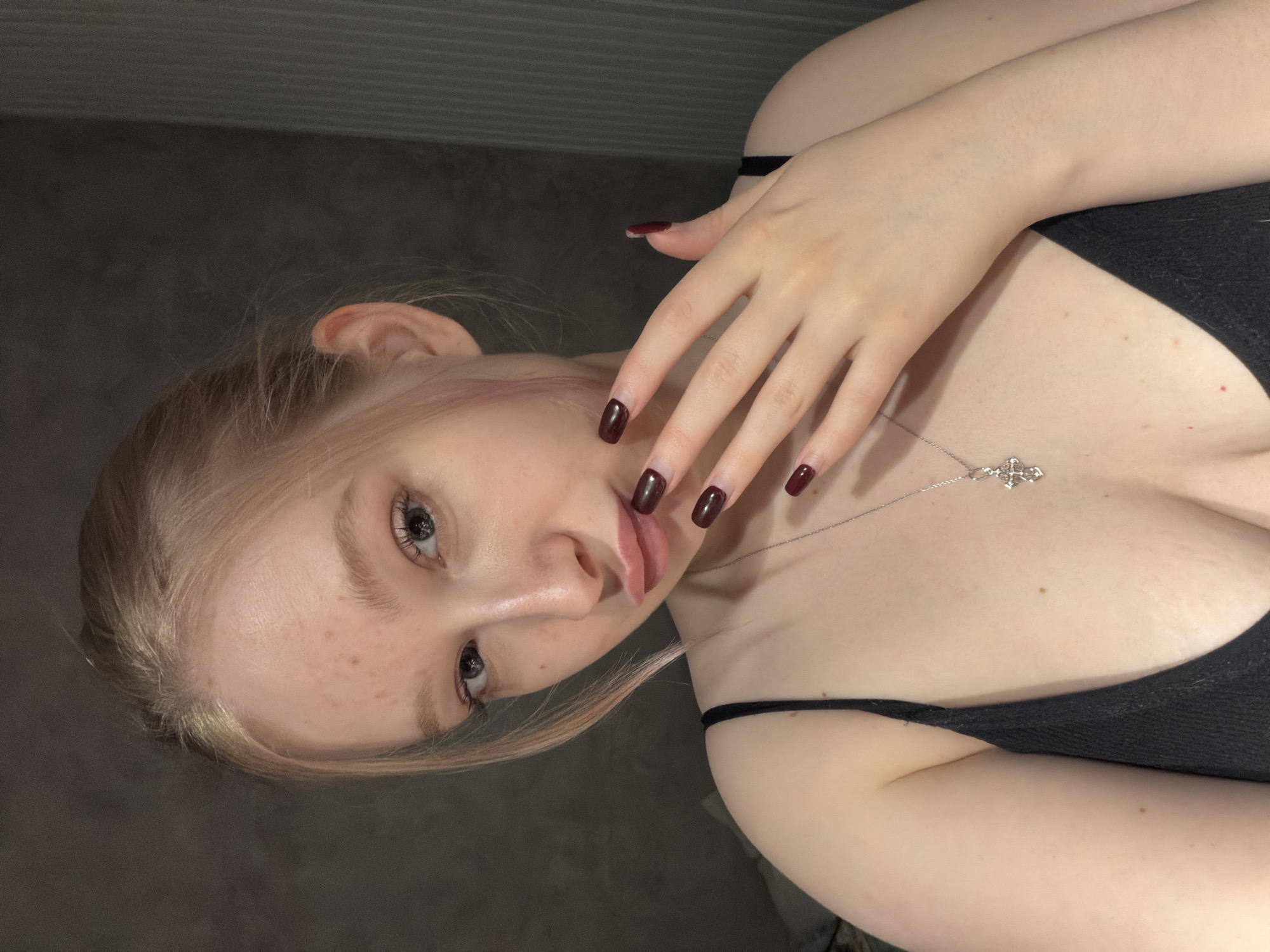 A fair-skinned person with blonde hair and visible freckles is angled towards the camera, with their head tilted towards their left shoulder. They are wearing a black, strappy top. A silver necklace with a cross pendant rests on their chest. Their hand, with dark red manicured nails, is partially covering their face. The background is a plain, gray wall. There is no explicit indication that the photograph was taken in the city of Rossiyskiy, Russia.