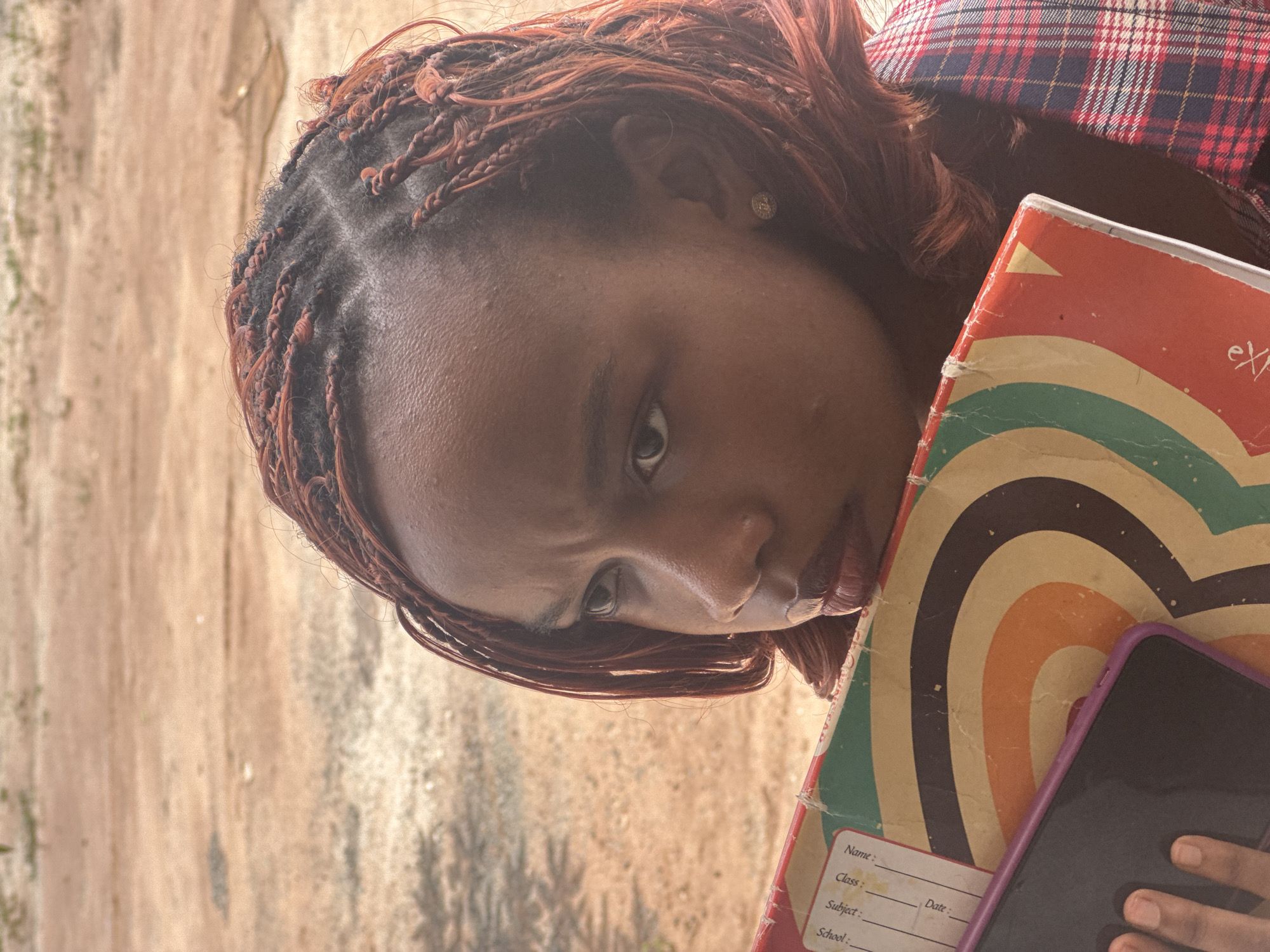 In Ihiagwa, Nigeria, a young Black woman with reddish-brown braided hair is pictured in a close-up shot, her face turned to her left, looking downwards with a contemplative expression. She is holding a vintage notebook with a colorful, circular design against her chest. A portion of a purple smartphone is visible in the lower left corner, held by her hand. Her attire includes a red and blue plaid shirt. The background is a blurred expanse of sandy earth, suggesting an outdoor, possibly rural or less developed, urban setting.