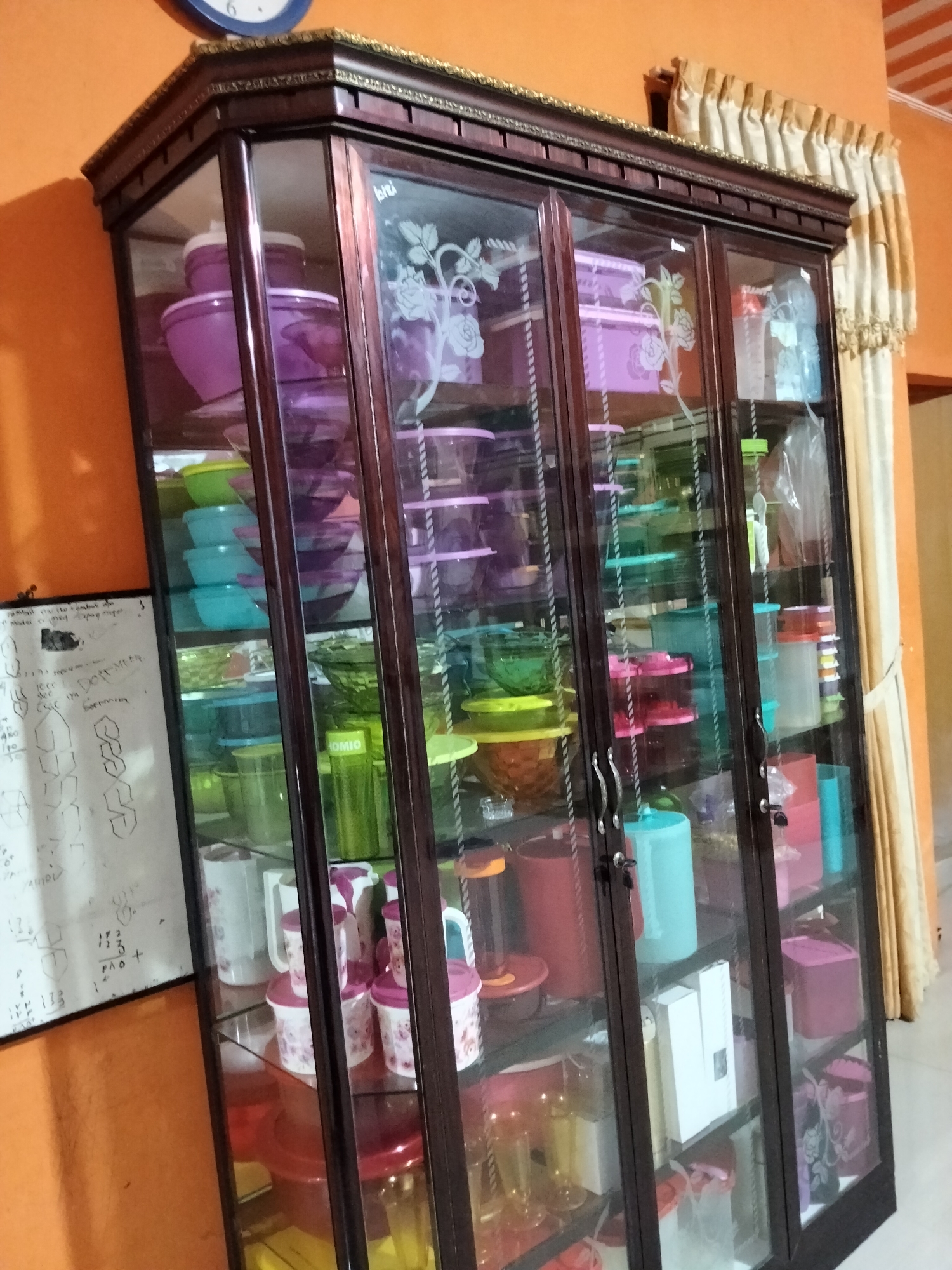 An indoor medium shot captures a dark wooden display cabinet with glass doors, filled with various plastic containers and kitchenware. The cabinet is situated against an orange wall, and to the right, a beige curtain with ruffled edges hangs. The cabinet's glass doors are decorated with etched floral patterns.
Inside the cabinet, on the top shelf, are purple plastic containers. Below these, on the second shelf, are stacked light blue and green plastic containers. The third shelf contains a variety of colored plastic bowls and containers, including green, red, and pink. On the fourth shelf, there are more containers, some red, some green, and a transparent bottle with a red lid. The bottom shelf holds more containers and bottles, including some pink ones. A white board with handwritten notes and diagrams is visible on the left side of the cabinet.
The scene appears to be a domestic interior, likely a kitchen or dining area. The overall impression is one of organized storage of household items. The lighting is ambient and appears to come from a light source above and to the left, casting some shadows. No people are present in the image.