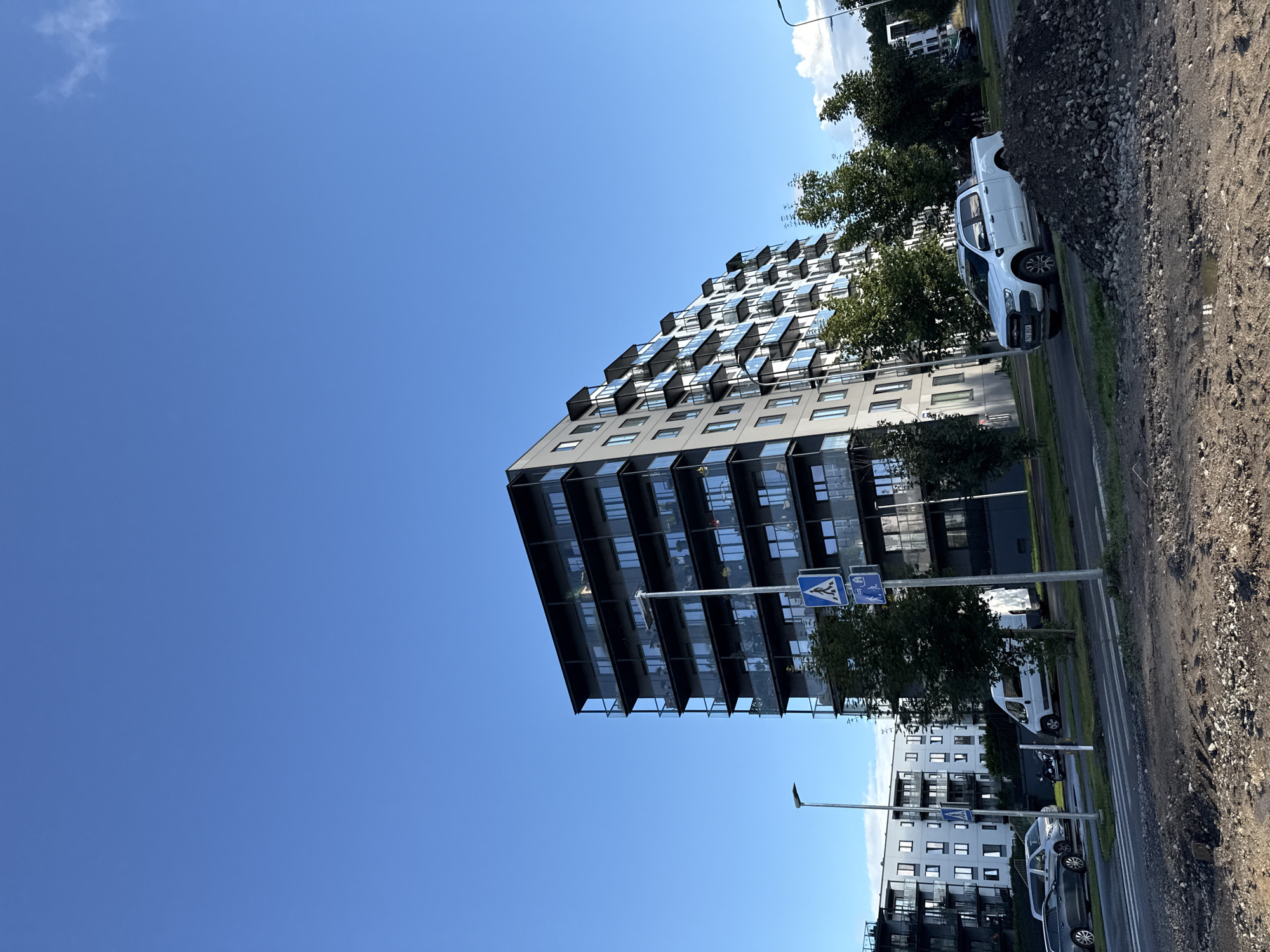 This is a street-level, slightly low-angle, exterior shot of a modern apartment building in Tallinn, Estonia. The building, with its dark frame and glass balconies, occupies the right half of the frame. In the foreground, there is a pile of gravel and dirt, suggesting ongoing construction. A white pickup truck is parked on the street in front of the pile. Further back, several other vehicles, including a white van, a dark sedan, and a silver SUV, are visible. Pedestrian crossing signs are present on poles along the sidewalk. Green trees are scattered around the building and along the street. The sky is a clear, bright blue with a few small clouds on the horizon.