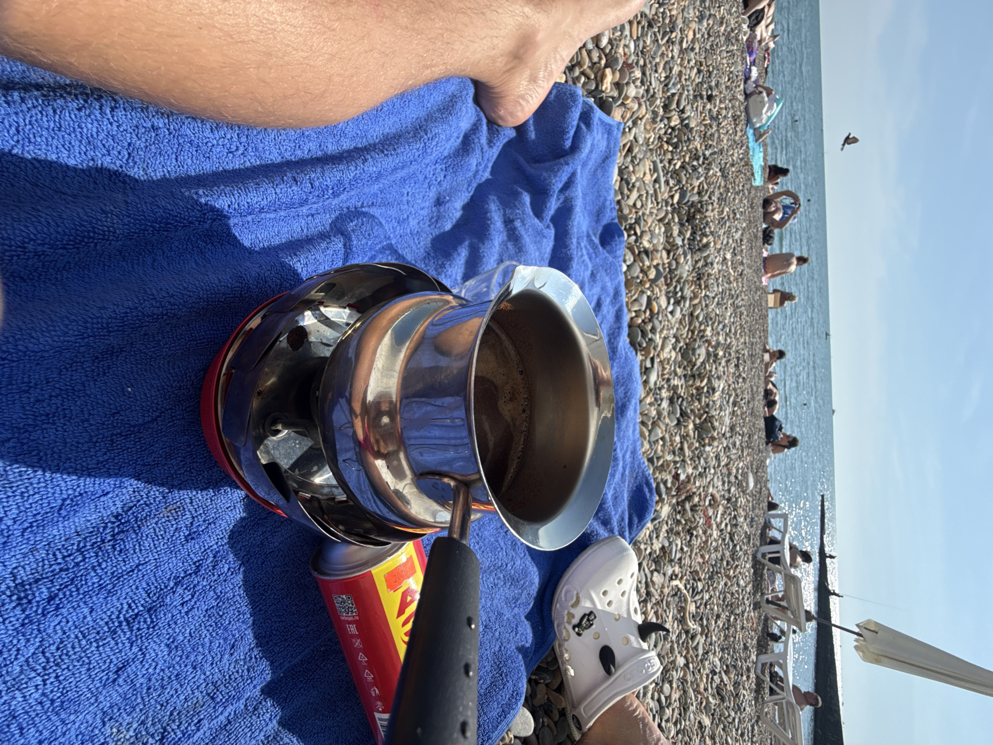 This is a close-up shot of a coffee pot sitting on a portable stove on a bright blue towel on a pebbly beach. In the background, the sea is visible with several people sunbathing and relaxing on loungers. The scene is set on a sunny day, likely in Sochi, Russia, judging by the architecture and atmosphere typical of the Black Sea coast. The focus is on the coffee-making setup, suggesting a leisurely moment by the sea.