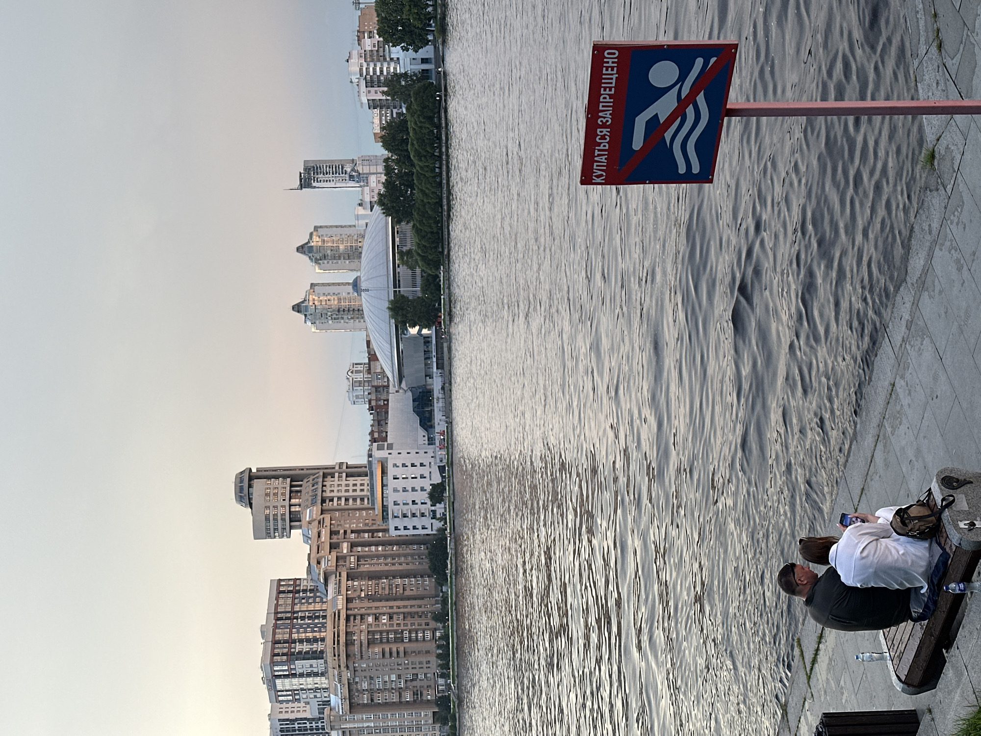 The image shows a waterfront scene in Yekaterinburg, Russia, likely on the Iset River. In the foreground, a man and a woman are sitting on a bench facing the water. The man is wearing a dark shirt and sunglasses, and the woman is wearing a white shirt. Both are looking at a smartphone. Next to them on the bench are two plastic bottles of water and a backpack. A "No Swimming" sign is prominently displayed in the mid-ground to the right, featuring a red circle with a diagonal line through a silhouette of a swimmer. The sign has Russian text that reads "КУПАТЬСЯ ЗАПРЕЩЕНО" which translates to "SWIMMING IS PROHIBITED." The river is choppy with visible ripples. In the background, a cityscape of Yekaterinburg unfolds with various multi-story buildings, including residential apartments and what appears to be a large domed structure. The sky is overcast with a soft, diffused light, suggesting either early morning or late evening.