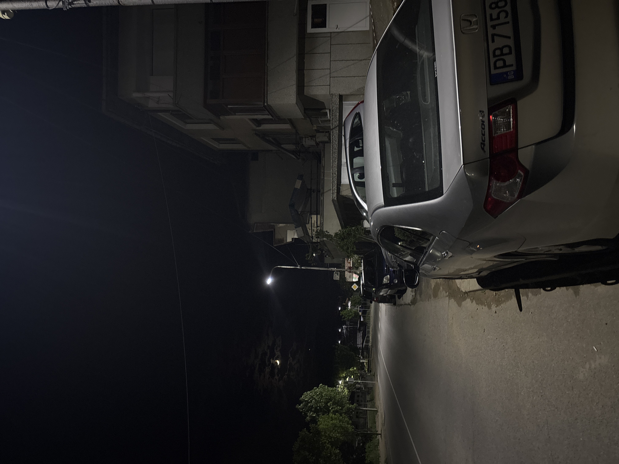 A nighttime street scene in Zlatograd, Bulgaria, shows a silver Honda Accord parked on the right side of the road. The car's license plate reads "PB 7158." Buildings line the left side of the street, and streetlights illuminate the scene. A crescent moon is visible in the night sky. The street is paved. Other vehicles are parked further down the street.