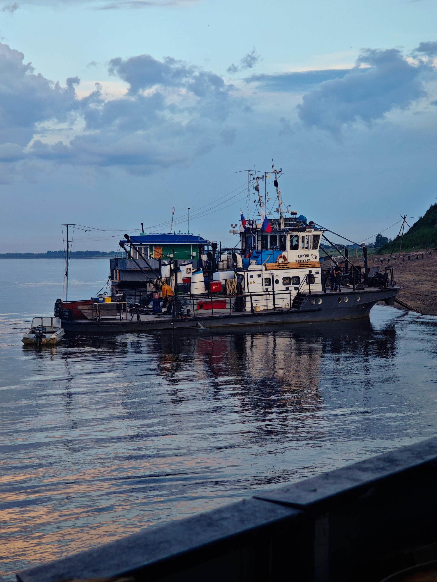 This media file shows a tugboat docked on a calm river. The tugboat, named "ГЕОРГИИ ПРОКОПЬЕВ" (Georgy Prokopyev), is gray with red and white accents. It has a multi-story superstructure, the upper section of which is painted blue. Russian flags are visible on the bridge. Two people are on the deck of the tugboat, one in a yellow jacket, and another in dark clothing. A small, gray dinghy with an outboard motor is tied to the side of the tugboat. The tugboat is grounded on a muddy bank, indicated by lines and structures on the shore, suggesting it might be undergoing maintenance or waiting for a tide.
The river water is calm, reflecting the sky and the boat. The sky is a mix of blue and gray clouds, with hints of pink and orange light, suggesting it is either early morning or late evening, likely during sunset or sunrise. The weather appears to be mild. The scene is set in Vorogovo, Russia, as indicated by the location context. The overall mood is peaceful and serene, with a sense of quiet activity on the boat. The foreground shows a dark, blurry railing, possibly from another vessel or a pier, further emphasizing the river setting.