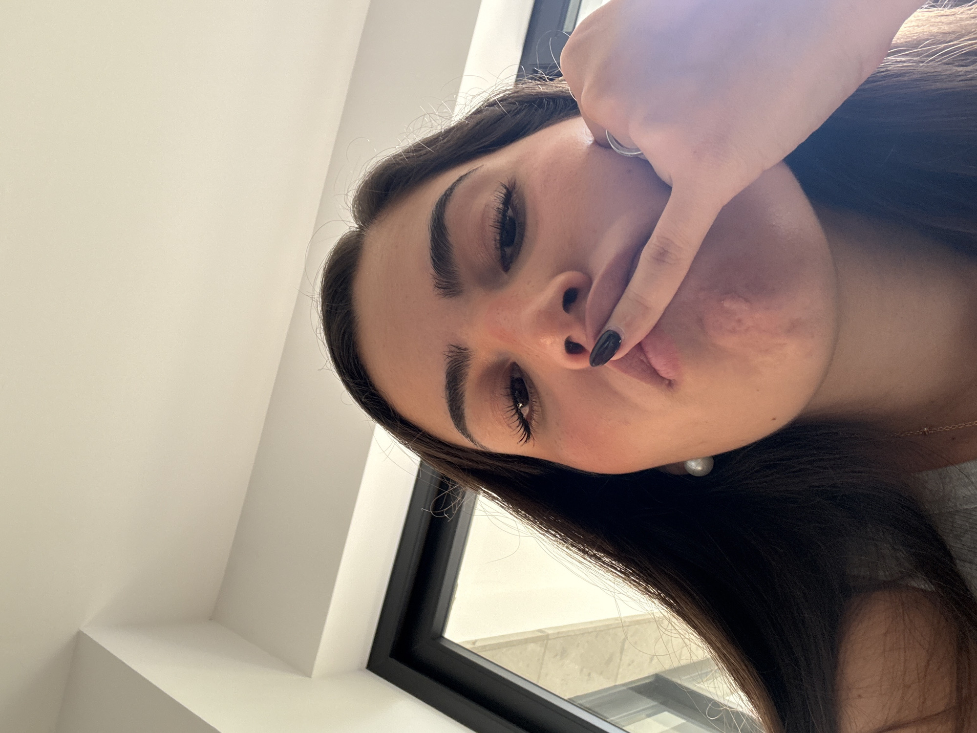 A woman with dark hair, dark eyebrows, and long eyelashes poses for a selfie indoors. She is holding a finger up to her lips. She wears a ring and has black nail polish on. A window with a black frame is visible behind her, as well as a white wall. The photo's location cannot be determined from the visual information provided.