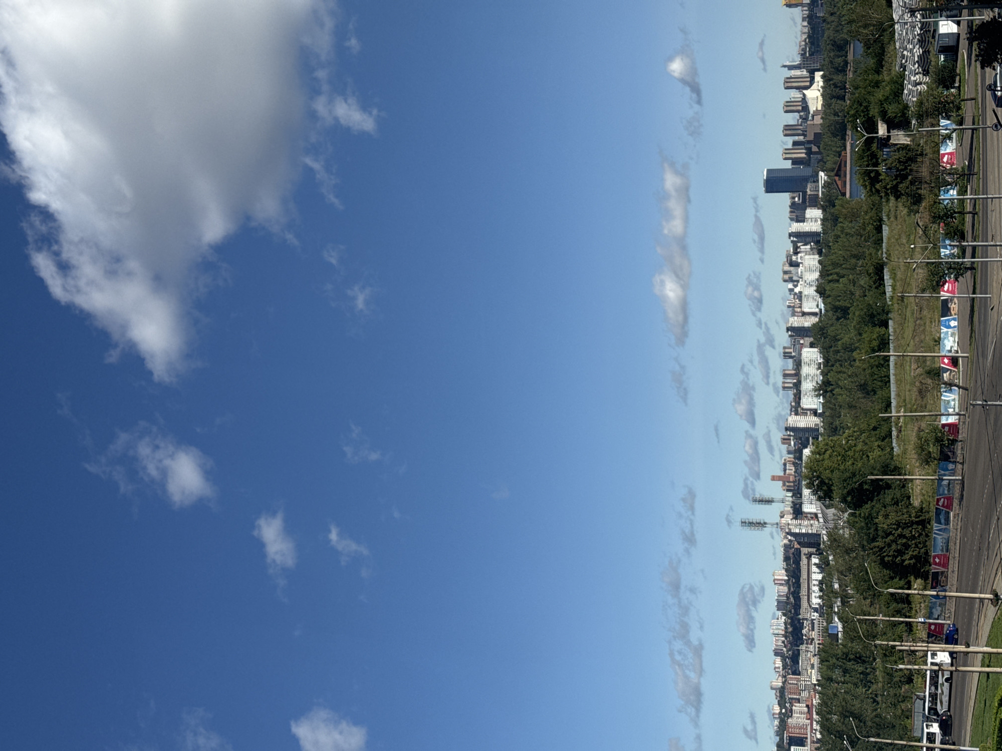 A wide-angle, eye-level shot captures a panoramic view of Krasnoyarsk, Russia. The sky is a vibrant blue, dotted with scattered white clouds. In the middle ground, a dense band of green trees forms a natural barrier between the foreground and the city skyline. Beyond the trees, the city unfolds, characterized by a mix of residential buildings and a few taller structures, including what appear to be communication towers. A prominent skyscraper stands out in the distant skyline. The foreground features a paved road with a sidewalk, and a few vehicles, including white buses and cars, are visible. Several lampposts line the roadside. In the far distance, hints of a body of water are discernible. The overall scene suggests an urban environment bordering a green, natural expanse on a clear day.