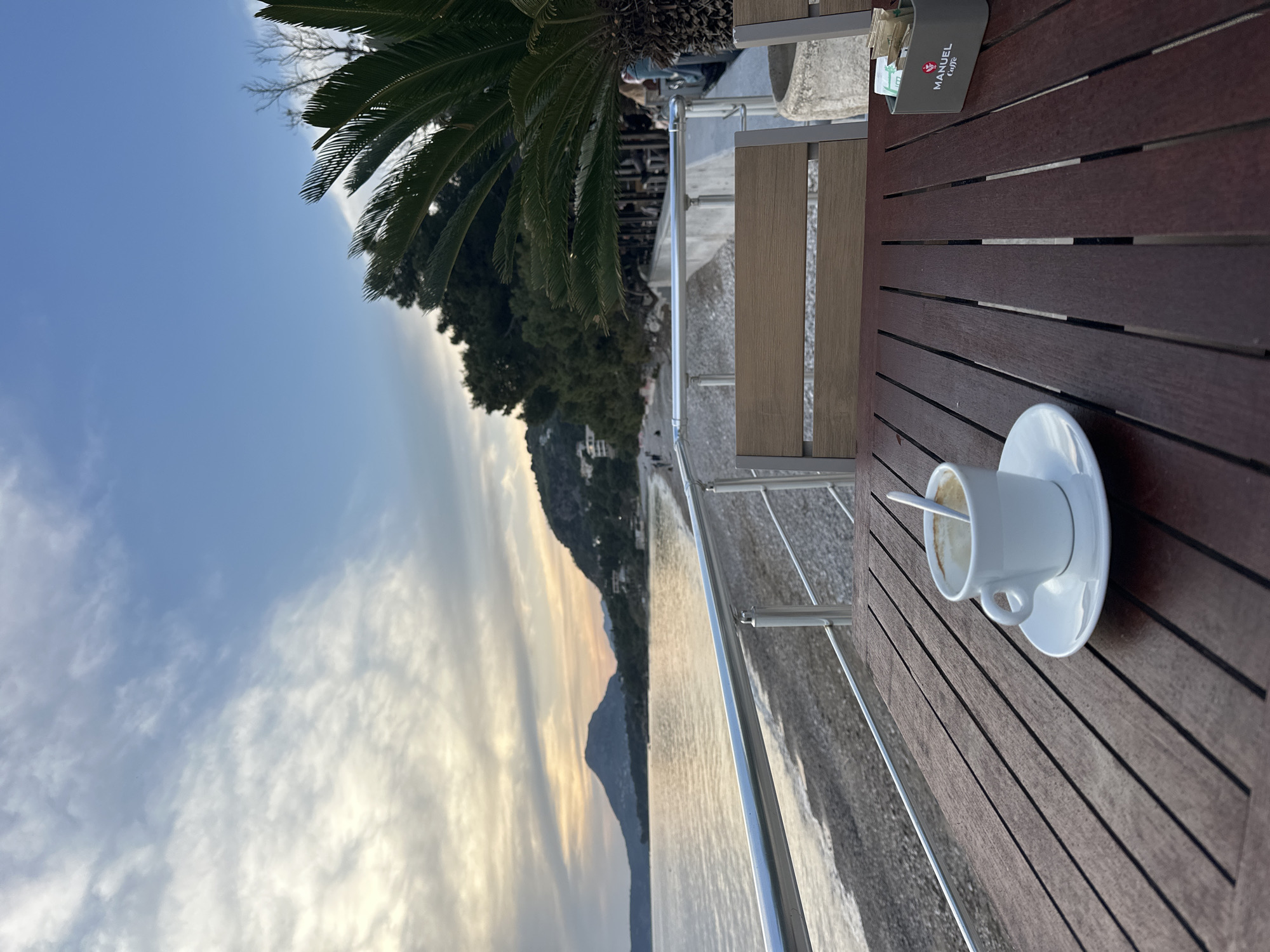The image shows a partially consumed cup of coffee on a dark wooden table at an outdoor cafe. The cafe overlooks the sea in Bar, Montenegro. A metal railing is visible, separating the cafe from a walkway or beach. In the background, there are trees, mountains, and the sky with clouds. A sugar packet holder with a logo saying "Manuel Caffe" is also on the table. A person sits in the background, along the side, with a part of the back of his head visible.