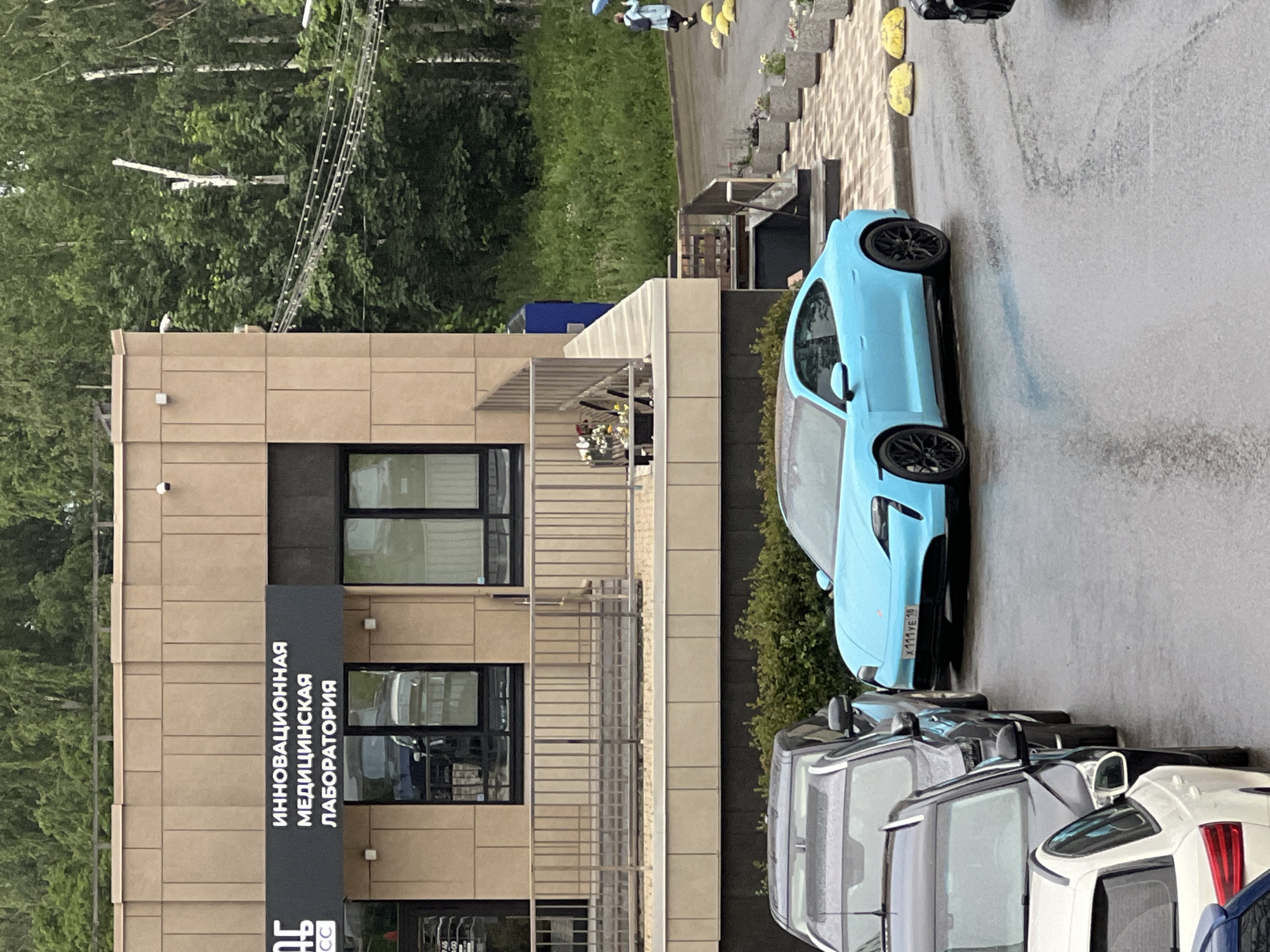 This media file captures a scene in Izhevsk, Russia, likely in the daytime. The weather appears overcast and damp, with wet asphalt visible on the ground. The primary focus is a bright blue electric car parked on a paved area next to a modern, beige-tiled building. The car has black rims and a license plate that reads "X 111 YE 19".
In front of the building, there is a dark sign with white text in Russian. The visible text includes "ИННОВАЦИОННАЯ МЕДИЦИНСКАЯ ЛАБОРАТОРИЯ", which translates to "INNOVATIVE MEDICAL LABORATORY". This suggests the building houses a medical facility.
Several other cars are parked in a line behind the blue electric car, mostly dark-colored. In the background, there are trees and a green grassy area, indicating a location with some natural surroundings. A partial view of a white car with red taillights is visible in the foreground to the right. The overall impression is an urban or semi-urban setting with a modern architectural element and parked vehicles.