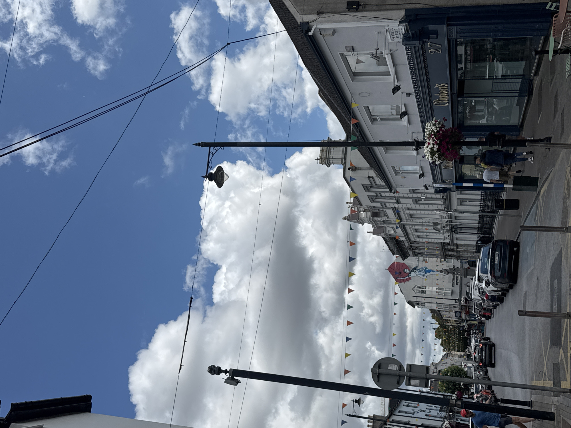 The image shows a street scene in Clonmel, Ireland, specifically Parnell Street. The sky is a bright blue with large, fluffy white clouds. Bunting in various colors is strung across the street. On the right side of the street, there is a building with a dark blue facade and a sign that reads "Clinton's". The building has large windows and is numbered 27. To the left of Clinton's, there are other buildings with grey and white facades. In the background, there is a mural of a woman holding a red umbrella and a statue. Several cars are parked along the street and some are driving. A few people are walking on the sidewalks. A tall lamppost with a light fixture stands on the left side of the street.