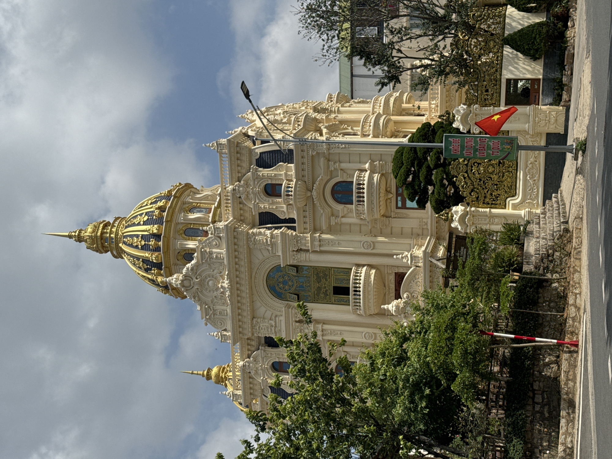 The image captures a grand building in Da Lat, Vietnam, likely a private residence or a hotel. The architectural style is heavily influenced by French colonial design, featuring ornate stucco details, decorative columns, arched windows, and a prominent dome with gold accents. A Vietnamese flag is visible at the entrance. Lush greenery surrounds the building, including trees and manicured shrubs. A street with visible pavement marks borders the property. A street light pole stands adjacent to the building. The sky is overcast with patches of blue visible through the clouds. A sign in Vietnamese is also visible at the entrance.
