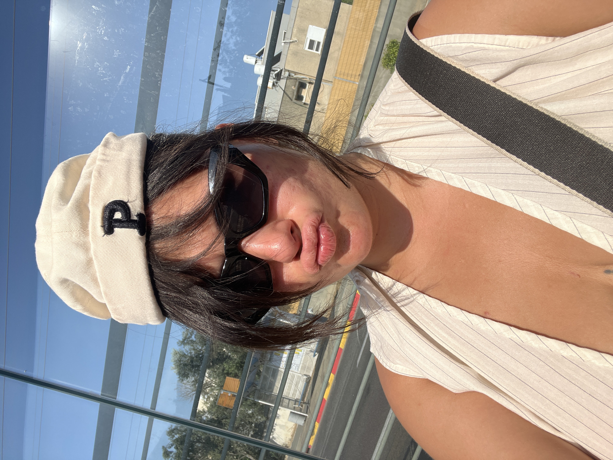 The image shows a woman with dark hair, wearing sunglasses, a white hat with a black "P" on it, and a pinstriped vest. She is puckering her lips for a selfie. She is standing in front of a glass bus shelter, with the city of Kiryat Motzkin, Israel visible in the background. The bus shelter has a metal frame and a translucent roof, and appears to be located near a street with a sidewalk. The background includes trees, buildings, and a bright blue sky. The lighting suggests it is daytime, with strong sunlight casting shadows.
