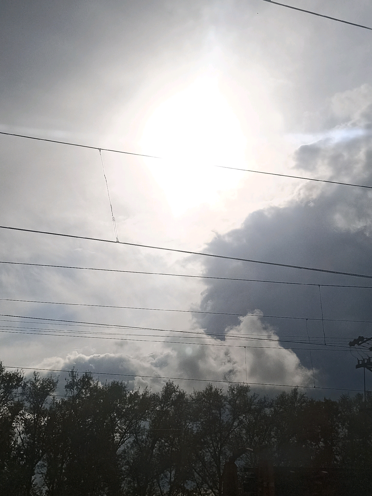 A low-angle, medium shot captures a portion of the sky above a treeline, viewed through a series of overhead wires. The sun is brightly visible, partially obscured by clouds which range from bright white to dark gray. Several horizontal wires crisscross the frame, with a few vertical supports visible on the right side. The lower portion of the image is dominated by the dark silhouettes of trees. The scene appears to be an outdoor urban environment, potentially a street or railway line in Kurundus, Russia, given the presence of overhead electrical wires. There are no people or identifiable subjects within the frame. The overall impression is of a cloudy, sunlit day.