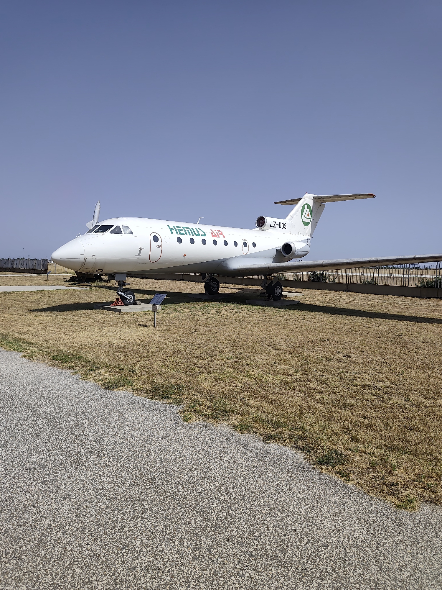 In Krumovo, Bulgaria, an abandoned Hemus Air Yak-40 aircraft with registration LZ-DOS is parked on a dry, grassy field under a clear blue sky. The aircraft is white with green lettering and a green logo on its tail fin. The foreground features a gravel path that curves away from the aircraft. The aircraft is stationary, with its landing gear deployed and supported by chocks. In the background, a chain-link fence runs horizontally across the scene. The environment appears arid, suggesting a dry climate.