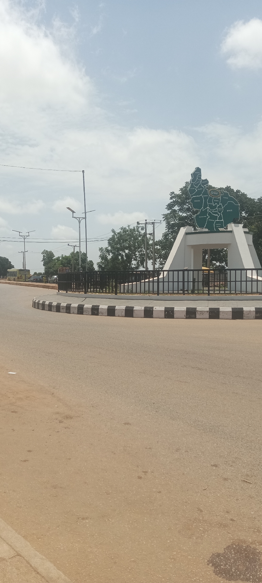 The image shows a roundabout in Kuru Dungus, Nigeria. The central feature of the roundabout is a white monument with a green map of the region on top. The map shows various districts with labels. The monument is enclosed by a black metal fence. The curb of the roundabout is painted with alternating black and white blocks. The road surface is light brown. Streetlights are visible in the background. Trees are present behind the monument. The sky is cloudy.