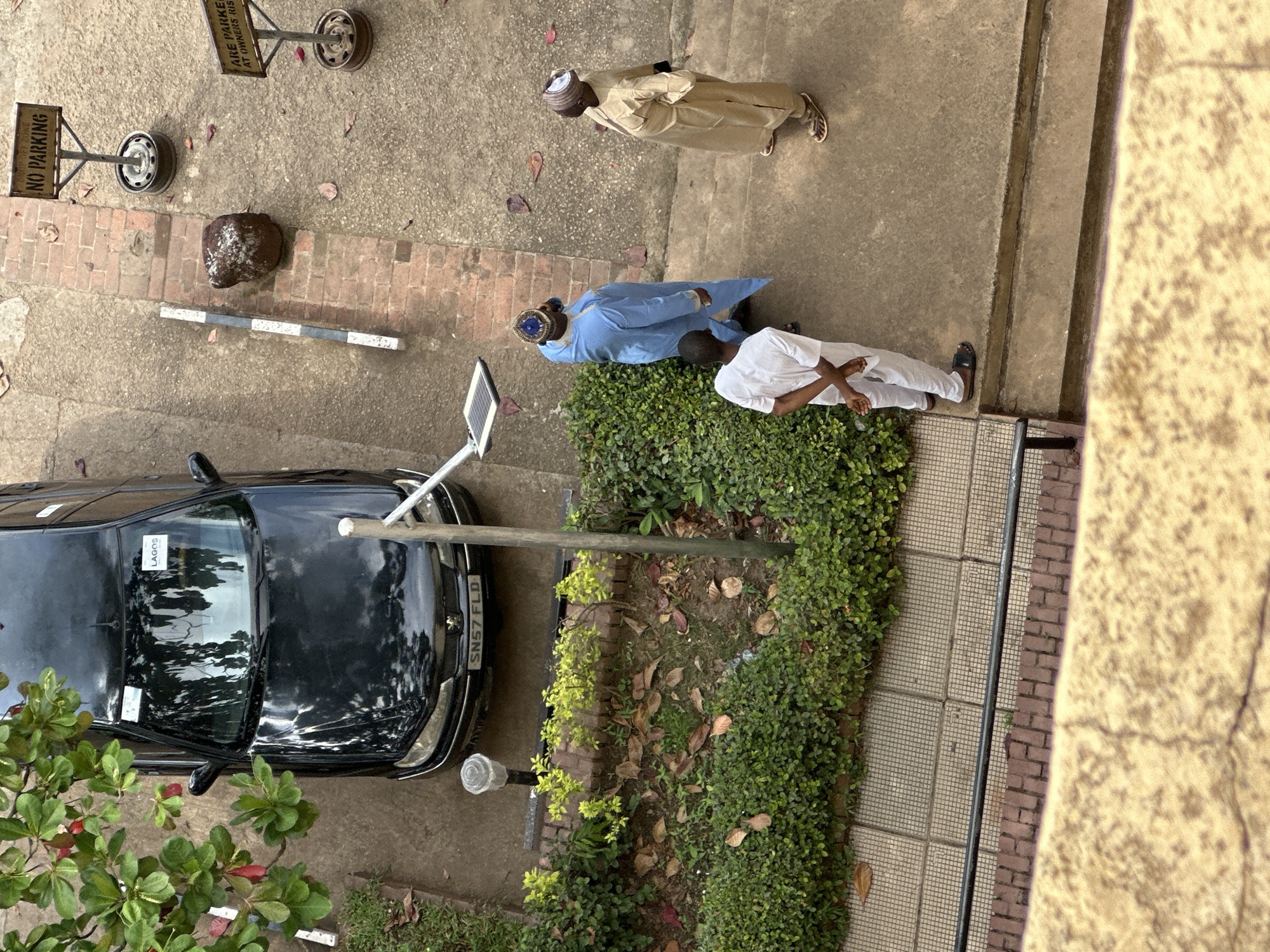 The image depicts an outdoor scene in the city of Shaba, Nigeria. A black Peugeot sedan is parked on a paved surface. In the foreground, three men are standing on concrete steps. Two of the men are wearing traditional Nigerian attire. One is in a blue robe and the other in a tan robe. The third man is in a white robe. To the left of the car, there are green bushes and trees. In the background, a "No Parking" sign is visible. The overall environment suggests a residential or administrative area.