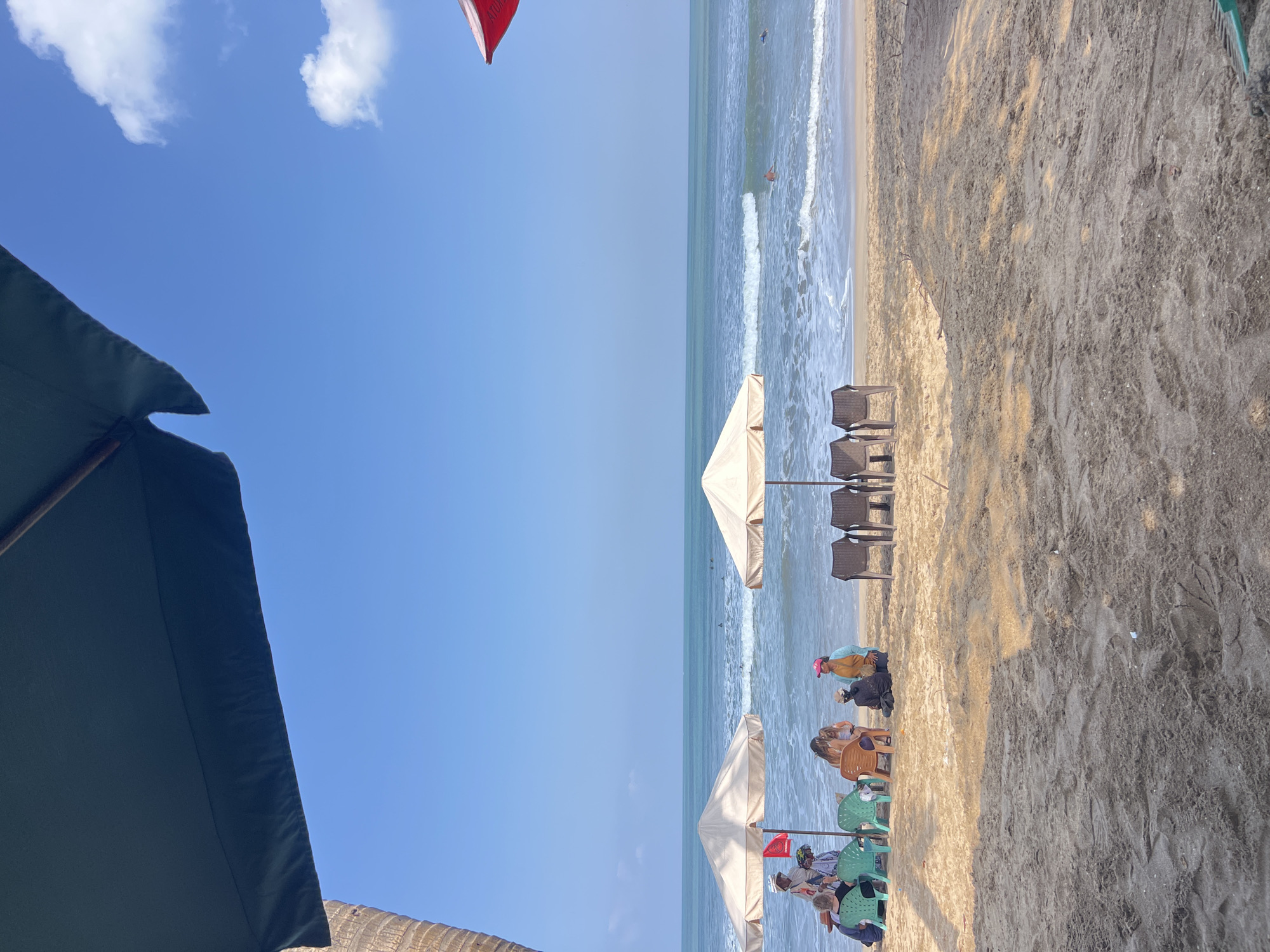 This image shows a serene beach scene in Kuta, Indonesia, under a clear blue sky with a few scattered clouds. The foreground is dominated by coarse, sandy beach, with tire tracks and scattered debris visible. In the mid-ground, several beach umbrellas are set up, providing shade for small groups of people seated on chairs or on the sand. The umbrellas are light-colored and appear to be made of fabric.
Two umbrellas are particularly prominent. The one on the left is near a small group of people standing and sitting. One person is holding a red flag. To the right of this group, under the same umbrella, a few people are seated. To the right of this, a second, larger umbrella is set up with a row of empty chairs beneath it. Further to the right, a partially visible red umbrella is in the upper right corner of the frame.
The ocean stretches out to the horizon, with gentle waves breaking on the shore. Several surfers are visible in the water, indicating moderate surfing conditions. A single person is also visible swimming or wading in the ocean. The water is a tranquil turquoise color. The overall impression is one of a peaceful day at the beach.