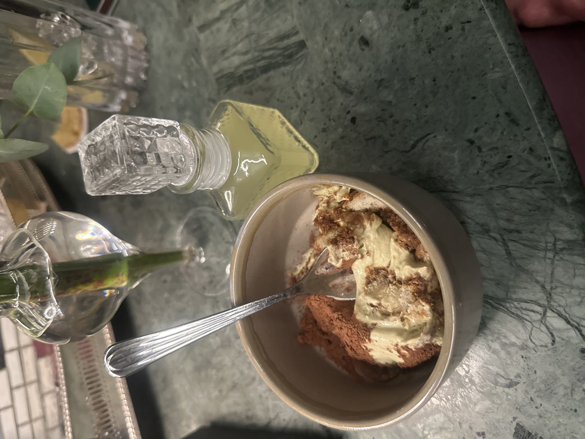 The image shows a close-up, overhead view of a dessert served in a beige bowl, set on a green marble surface. The dessert is layered with a light-colored cream and a dark, possibly chocolate-based, layer. A silver fork rests in the bowl.
Beside the bowl, there's a small glass bottle with a decorative stopper and a light-colored liquid. To the left, there is also what appears to be a glass vase with a long-stemmed flower.
The specific location is not discernible from the image. No information about people, specific actions, or interactions is present.
