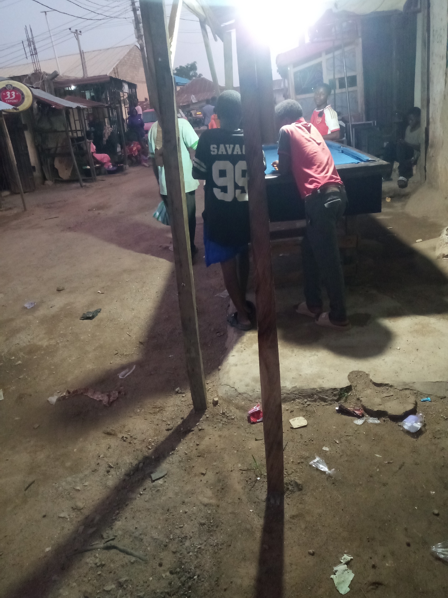 The scene is an outdoor pool hall in Jos, Nigeria. Two men are playing pool, one wearing a red shirt and the other wearing a black jersey with the number 99 and "SAVAI" on the back. Another person in a green shirt stands nearby. The pool table is illuminated by a bright light. In the background are market stalls, other people, and buildings. The ground is dirt with debris scattered around. Two wooden poles frame the scene.