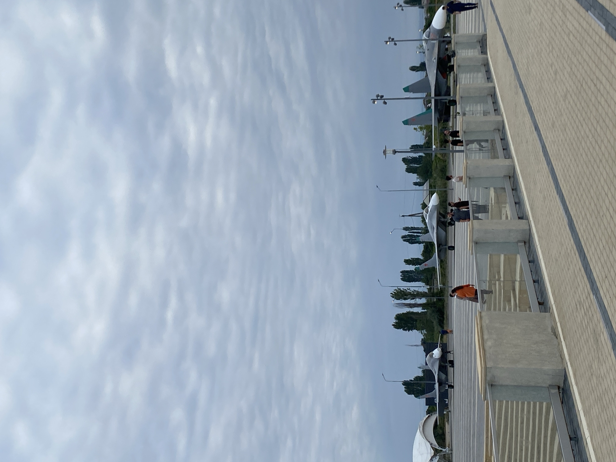 The image captures an outdoor scene with several military jets parked on a tarmac. The sky is overcast with gray clouds. In the foreground, there is a paved walkway and a low concrete barrier with a glass railing. Behind the barrier, there are stairs leading down. Several people are visible, some standing near the barrier and others walking on the tarmac. In the middle ground, two fighter jets are prominently displayed. One jet is gray with a white nose cone, and the other is a darker gray with teal accents on its tail fins. Further back, more trees and structures are visible, suggesting an airfield or an aviation museum. The location is identified as Arablyar, Russia.