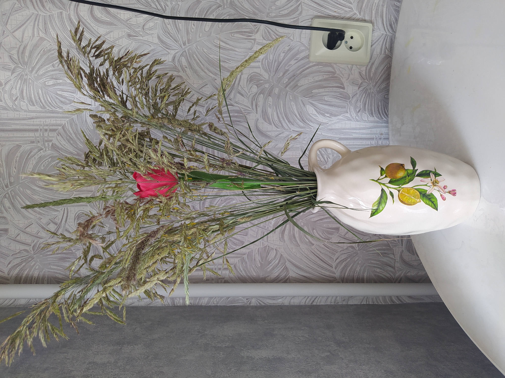 A still life photograph taken indoors displays a white ceramic vase with a lemon and blossom design. The vase, positioned on a white table, holds a bouquet of dried grass and a single bright pink tulip. Behind the vase, a light-colored wall with a leaf-patterned wallpaper is visible. To the right of the vase, a white electrical outlet with a black cord plugged in is mounted on the wall. A white vertical pipe is also present on the left side of the image. The overall scene suggests a domestic setting. The location is Gusev, Russia.