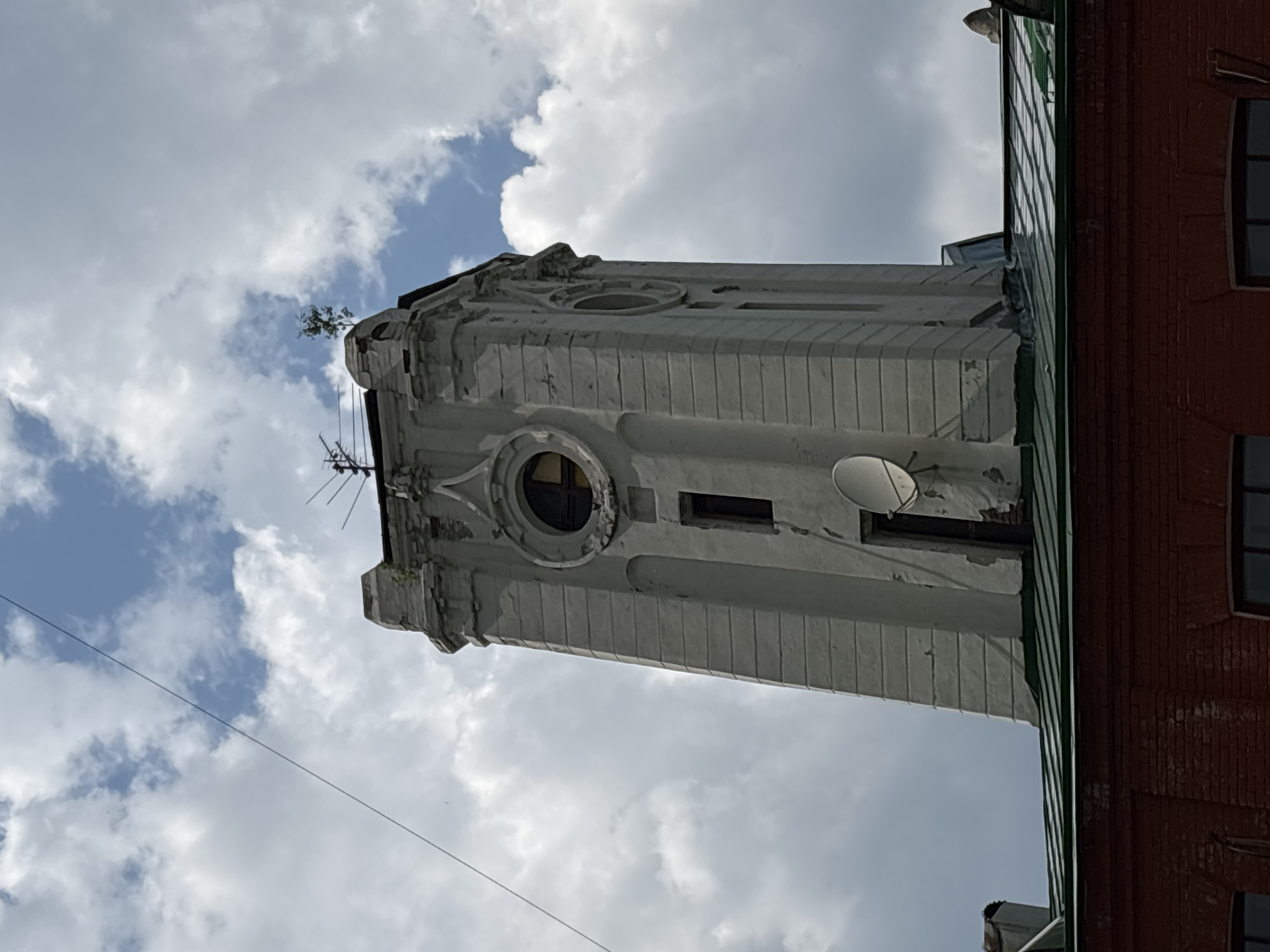 A tall, weathered, pale gray tower with a square base and ornate detailing stands against a cloudy sky. The tower has several windows, including a large, circular one near the top with yellow panes. A small satellite dish is attached to the side of the tower. A thin wire stretches across the upper left portion of the image, originating from above and to the left of the tower. The tower is situated above a red brick building with a green metal roof. The overall impression is of an old, possibly industrial or civic, structure. The image is of the clock tower of the Kirovsky District Administration Building in Novosibirsk, Russia.