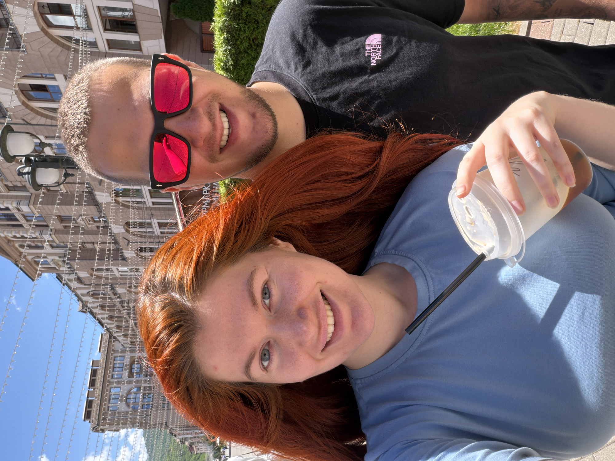 In the city of Estosadok, Russia, a close-up, waist-up outdoor selfie captures a young couple on a sunny day. The woman, positioned on the left, has long, vibrant red hair that cascades over her shoulders. She is wearing a light blue t-shirt and is holding a clear plastic cup with a dark straw, seemingly mid-sip. Her face is turned towards the camera, and she has a bright, genuine smile, with her blue eyes visible and a scattering of freckles across her nose and cheeks. The man, on the right, is wearing a black t-shirt with the "The North Face" logo subtly visible on his left chest. He has short, light brown hair and is wearing large, black-framed sunglasses with bright red lenses that reflect the sunlight. His face is turned towards the camera, and he is also smiling. In the background, the architecture of Estosadok is visible, with ornate buildings featuring arched windows and balconies, suggesting a charming, possibly historic, urban setting. Strings of decorative lights or garlands are strung across the street overhead, adding a festive touch to the scene. The lighting indicates it is midday, with clear blue skies and bright sunshine casting shadows on the ground.