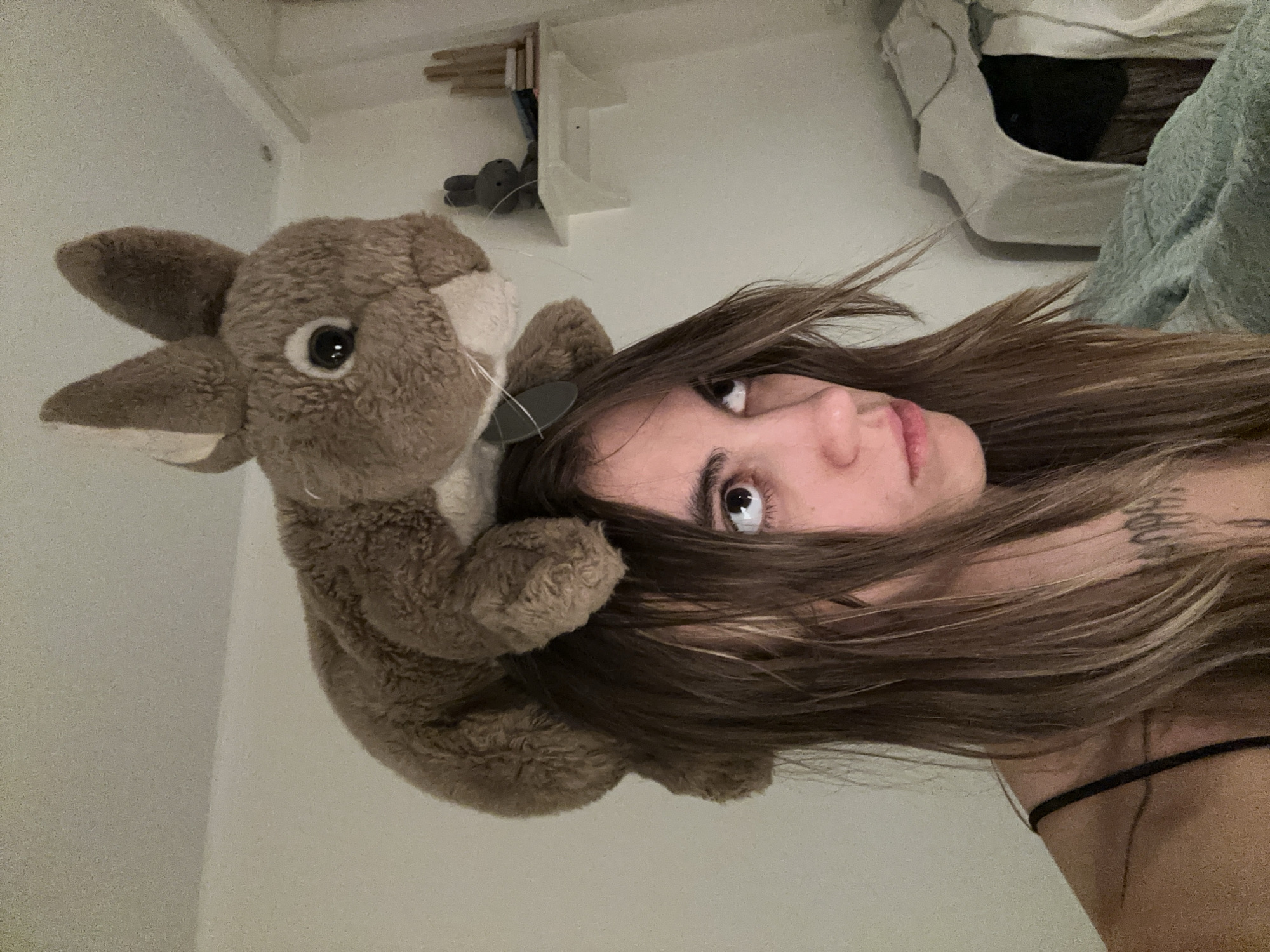 A person with long, light brown hair is looking to their right, with a stuffed rabbit on top of their head. The person is wearing a black strap top, and has tattoos on their chest and shoulder. The background appears to be a bedroom, with a shelf on the wall holding a small gray stuffed bunny and some books. The overall lighting is dim, suggesting indoor lighting. The image does not contain enough information to determine the location in Moscow, Russia.